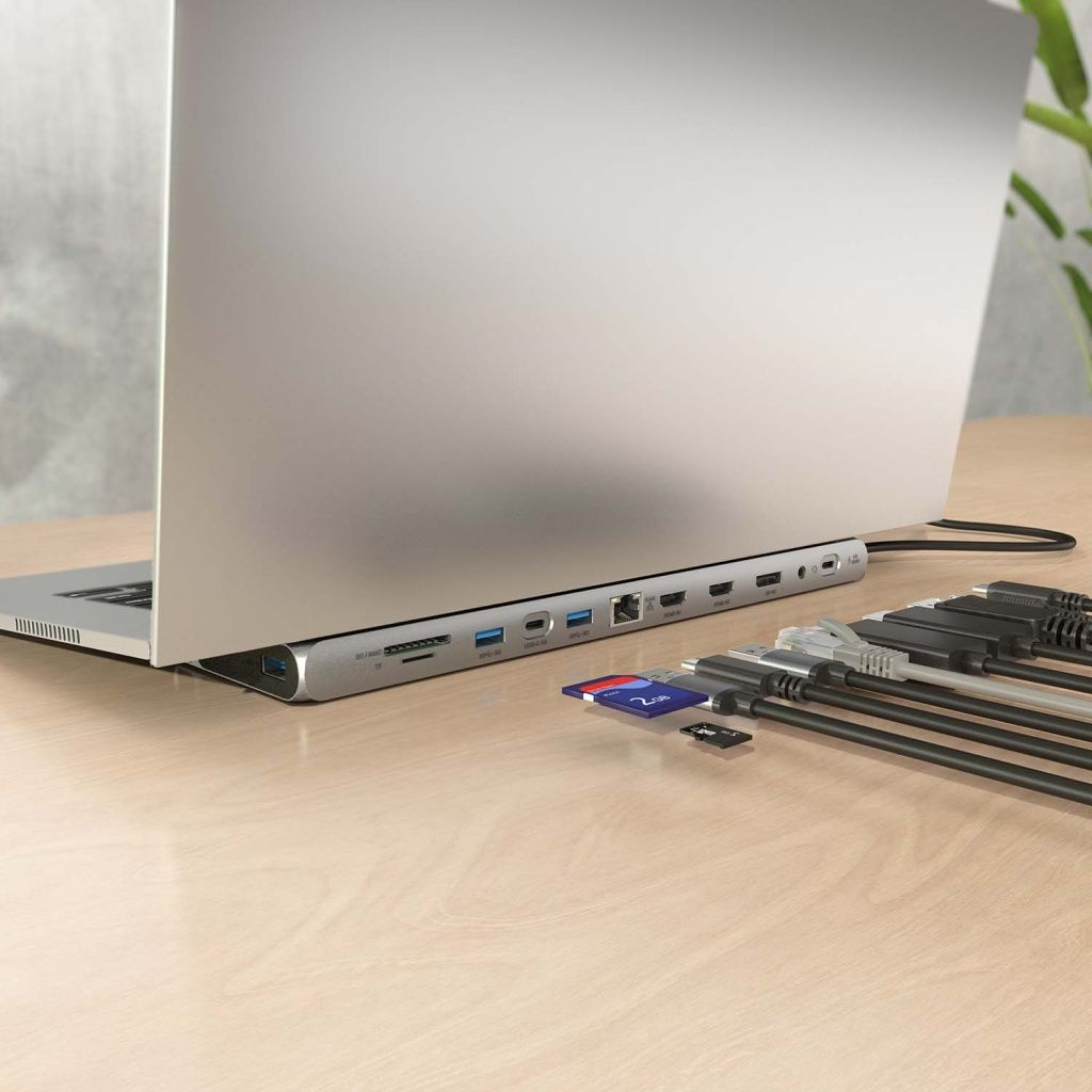 Un portátil conectado a un hub USB plateado con varios puertos. Cables y tarjetas SD conectadas. Fondo: mesa de madera y planta.