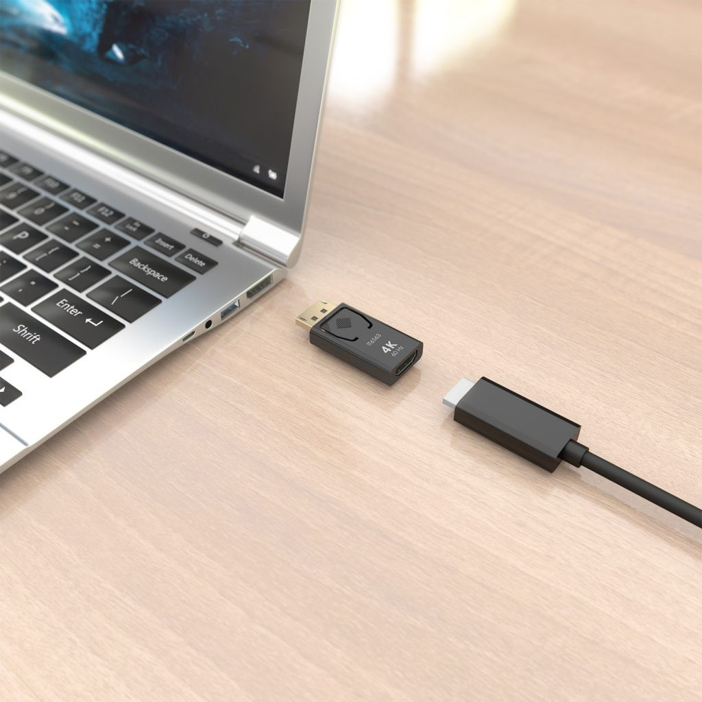 Ordenador portátil con un adaptador HDMI negro y un cable sobre una mesa de madera clara. El adaptador está etiquetado como 4K 60Hz.
