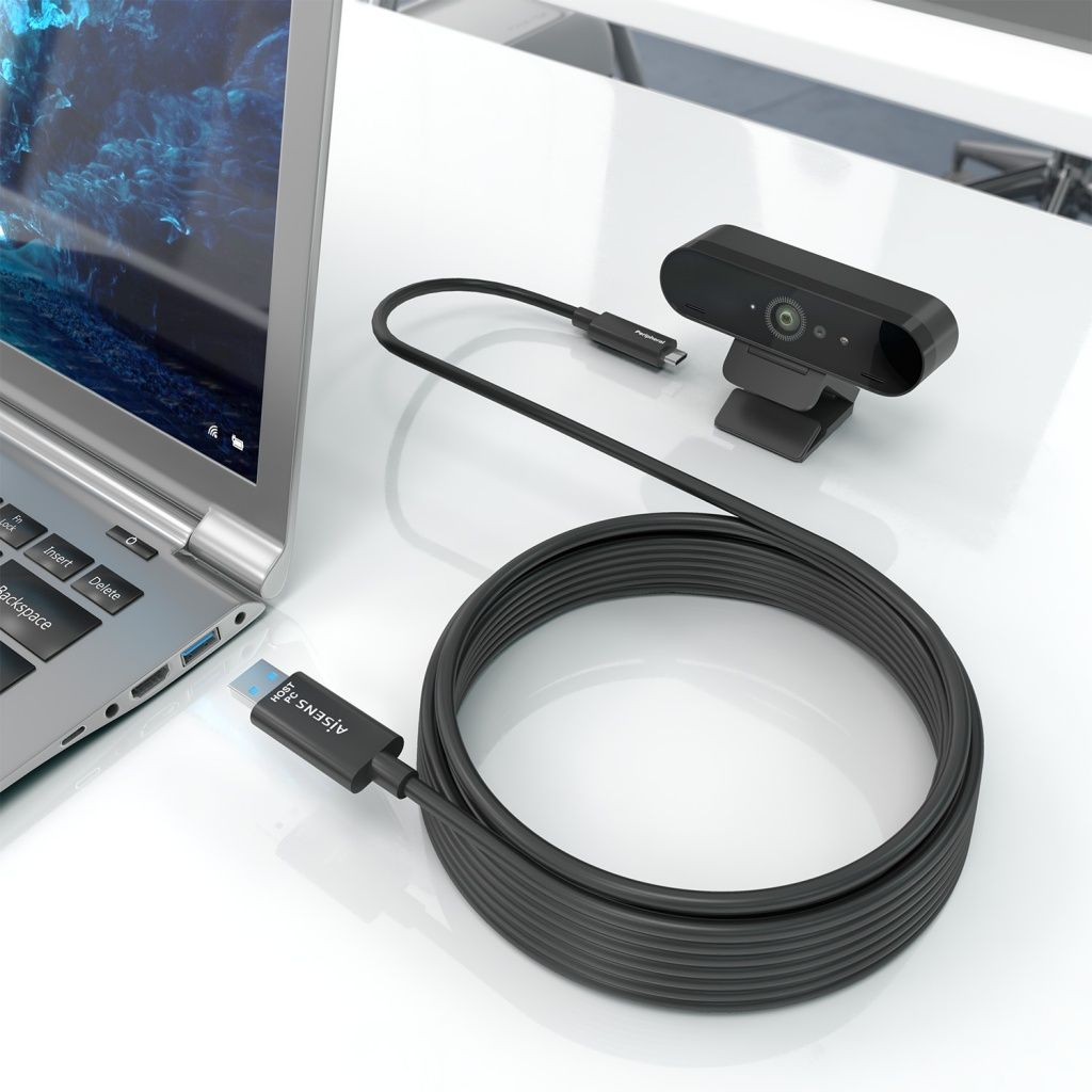 Una cámara web negra con un cable USB está junto a una computadora portátil. El cable está enrollado y conectado a la computadora portátil.