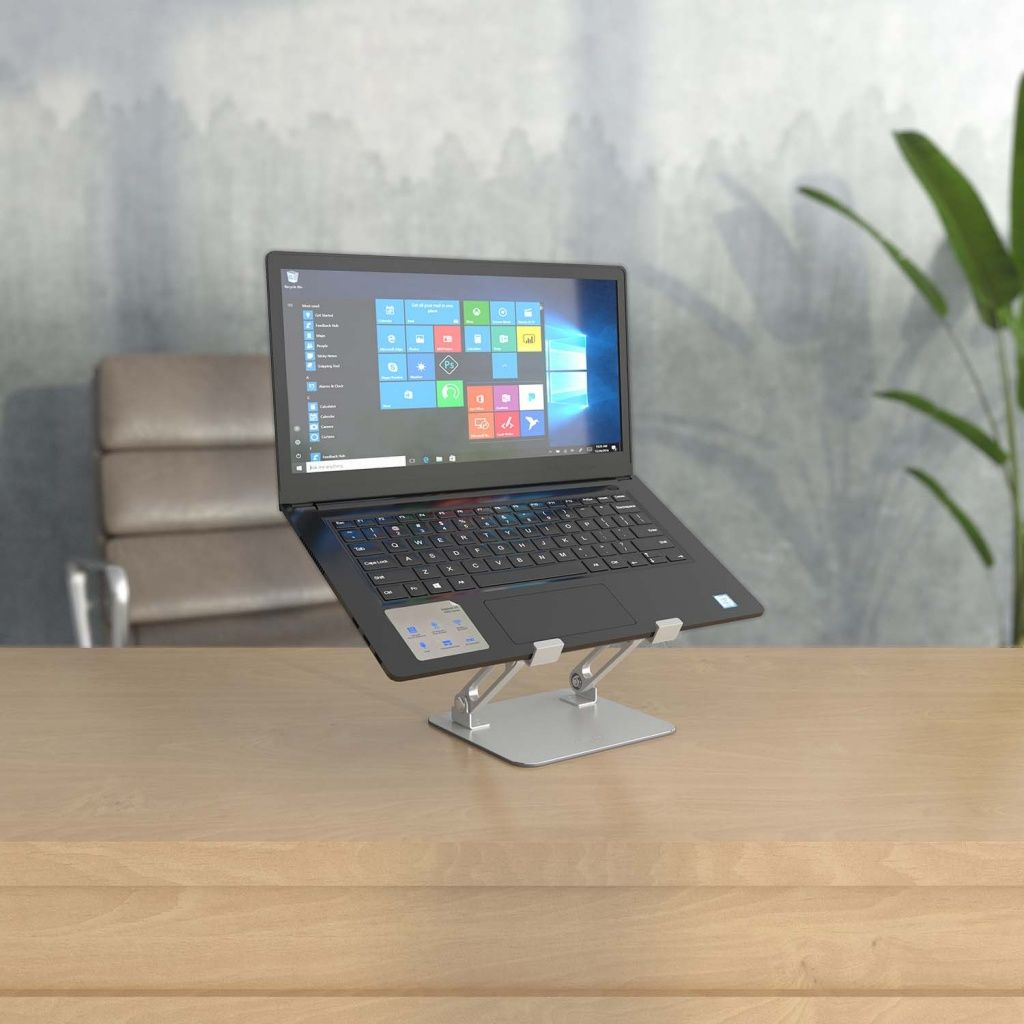 Een zwarte laptop staat open op een zilveren standaard op een houten bureau. Het scherm toont het Windows startmenu. Een stoel en plant staan op de achtergrond.
