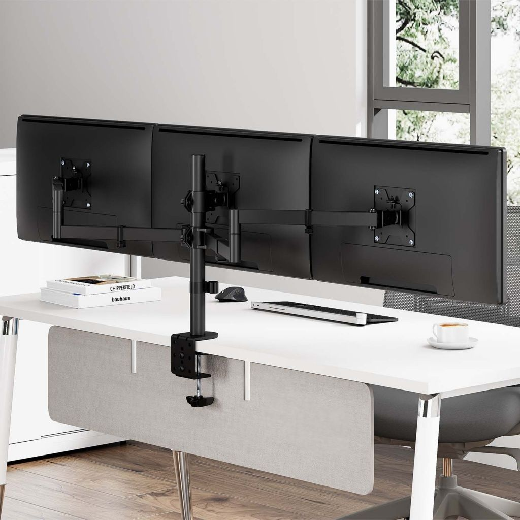Tres monitores de ordenador negros sobre un escritorio blanco con un soporte. Libros, teclado, ratón y taza de café están sobre el escritorio.