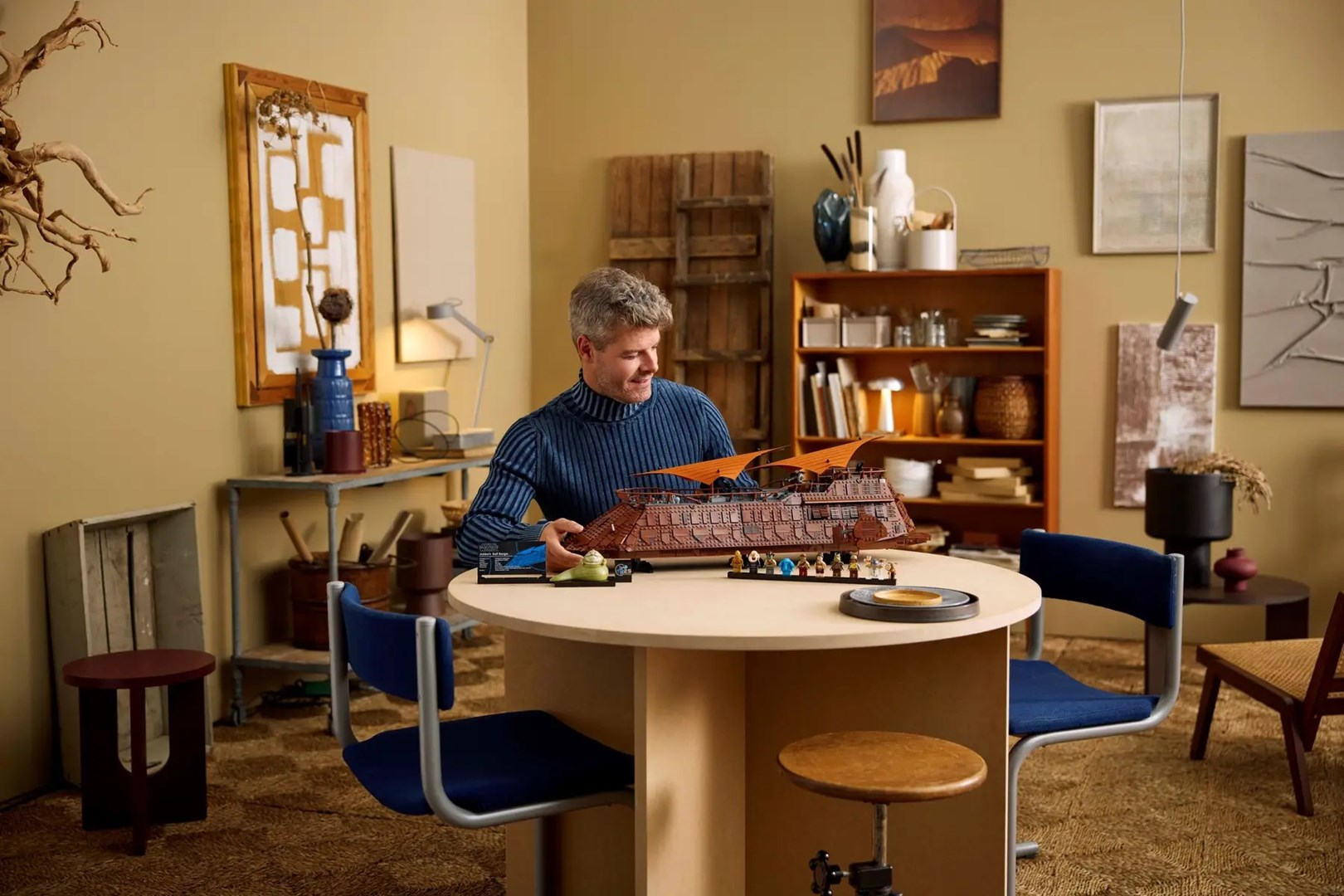 Mężczyzna buduje model Lego statku przy stole. Pokój ma drewniane meble i sztukę na ścianie.