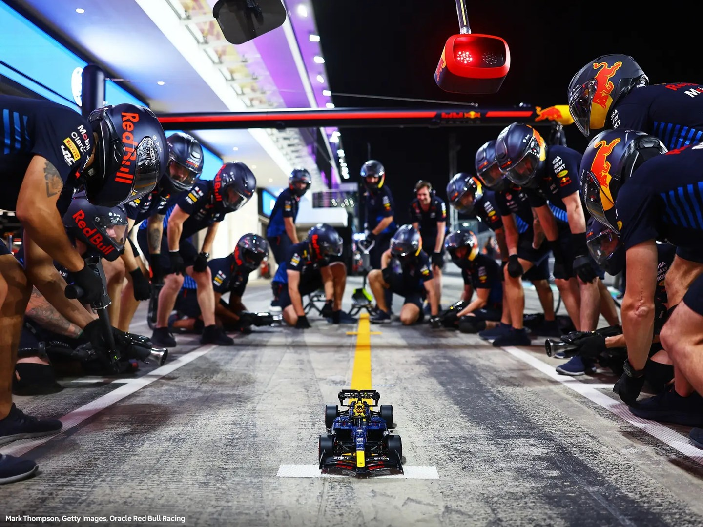 Samochód Formuły 1 otoczony przez zespół podczas postoju w boksie. Członkowie zespołu noszą kaski i pracują przy samochodzie. Scena ma miejsce w alei serwisowej.