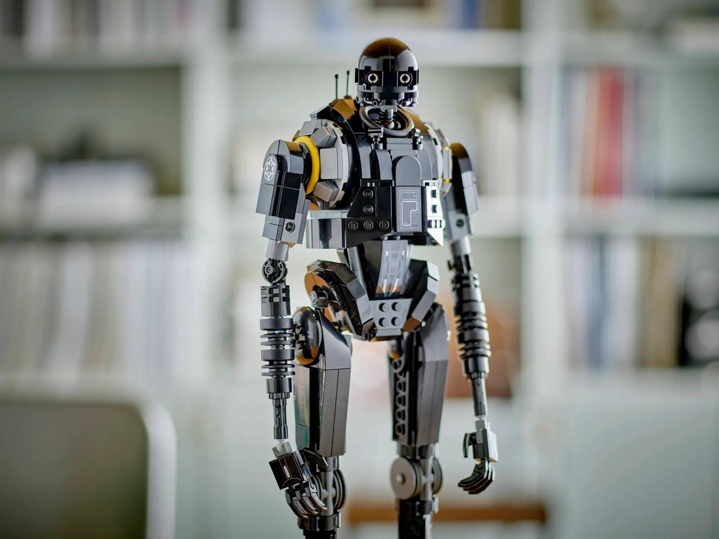 Czarna figurka robota Lego K-2SO stoi na tle rozmytej półki z książkami. Ma czarną głowę, tułów, ramiona i nogi z żółtymi akcentami.