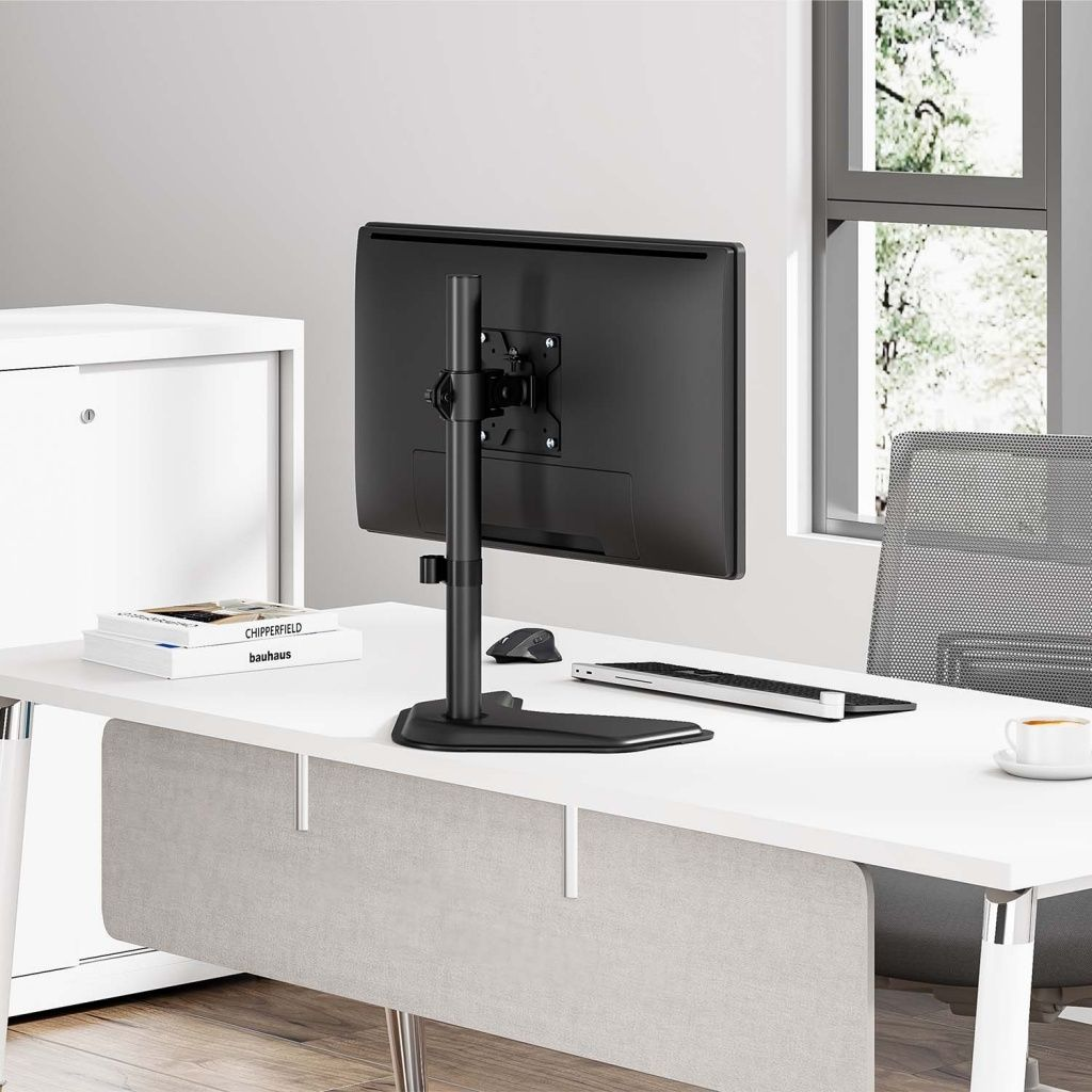 Un monitor negro sobre un soporte está sobre un escritorio blanco. Un teclado, un ratón y una taza también están sobre el escritorio. Una ventana está en el fondo.