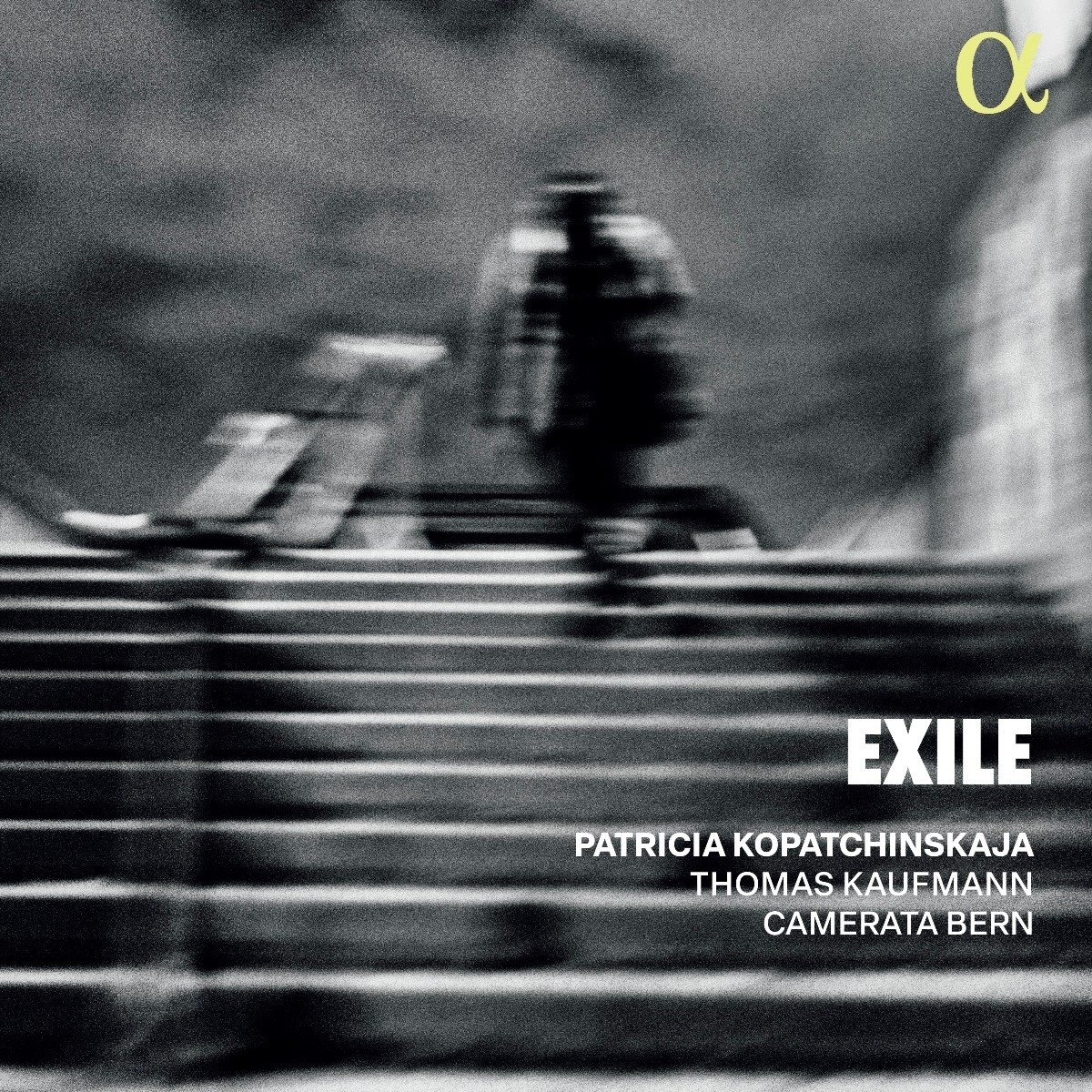 Immagine monocromatica di una persona sfocata che sale le scale. Titolo: EXILE. Nomi: Patricia Kopatchinskaja, Thomas Kaufmann, Camerata Bern.