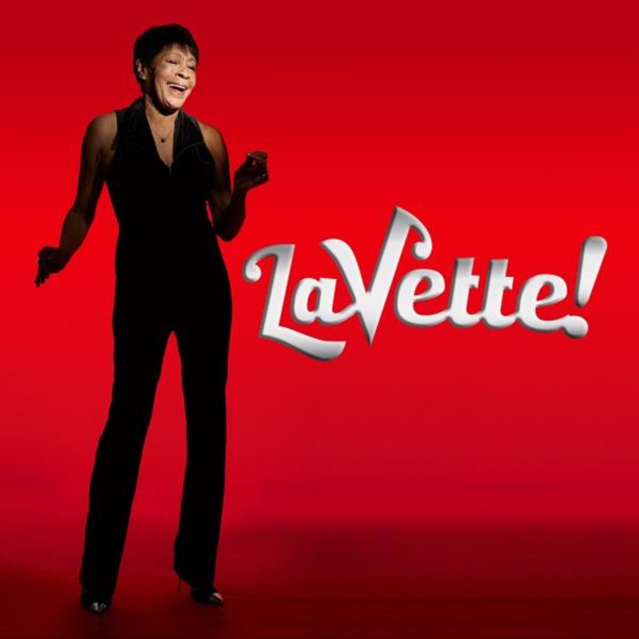 Een vrouw in zwarte outfit lacht, houdt een voorwerp vast, met rode achtergrond en tekst "LaVette!".