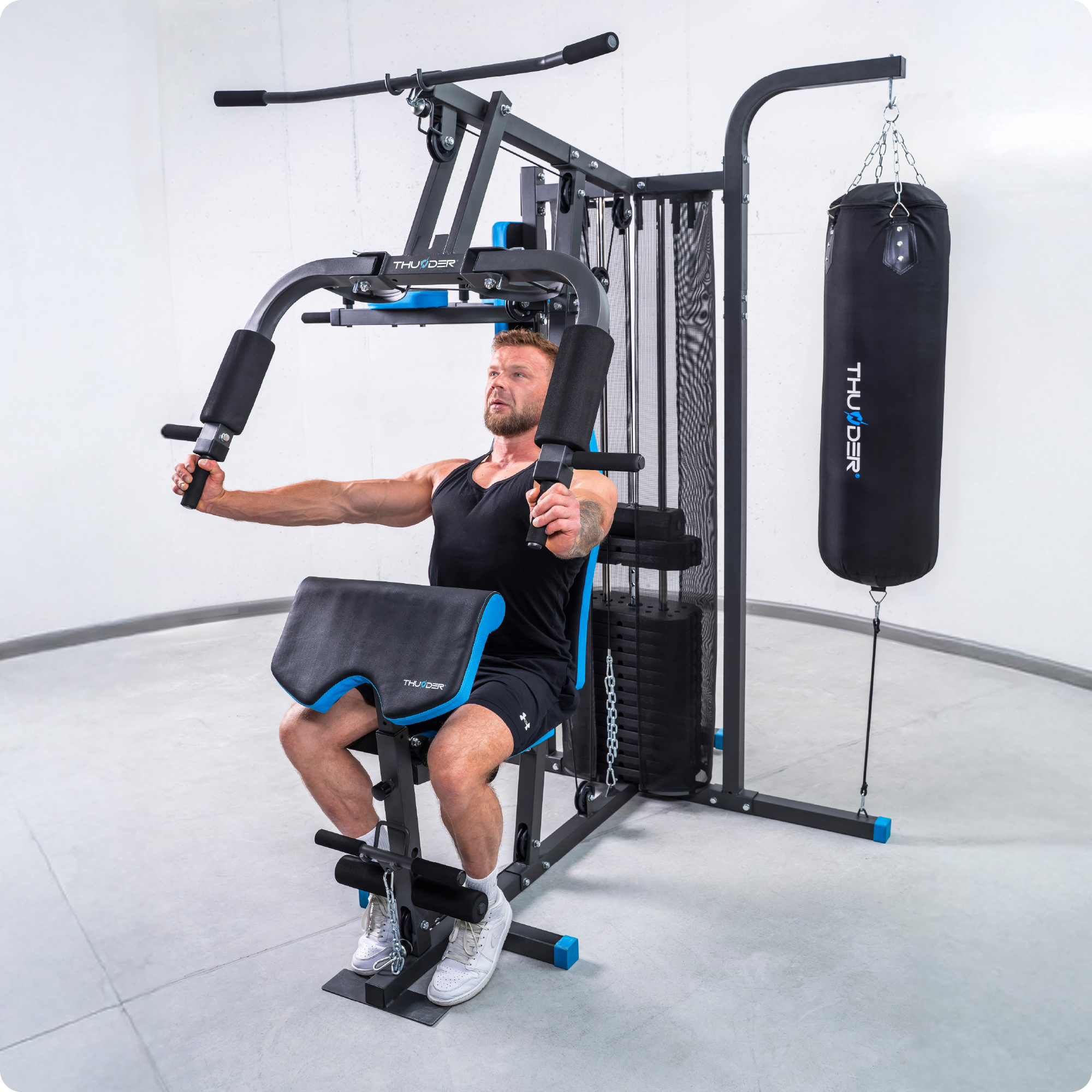 Mężczyzna korzysta z multi-gimnastyki. Siłownia ma czarne i niebieskie akcenty. W tle wisi worek treningowy. Podłoga jest jasnoszara.