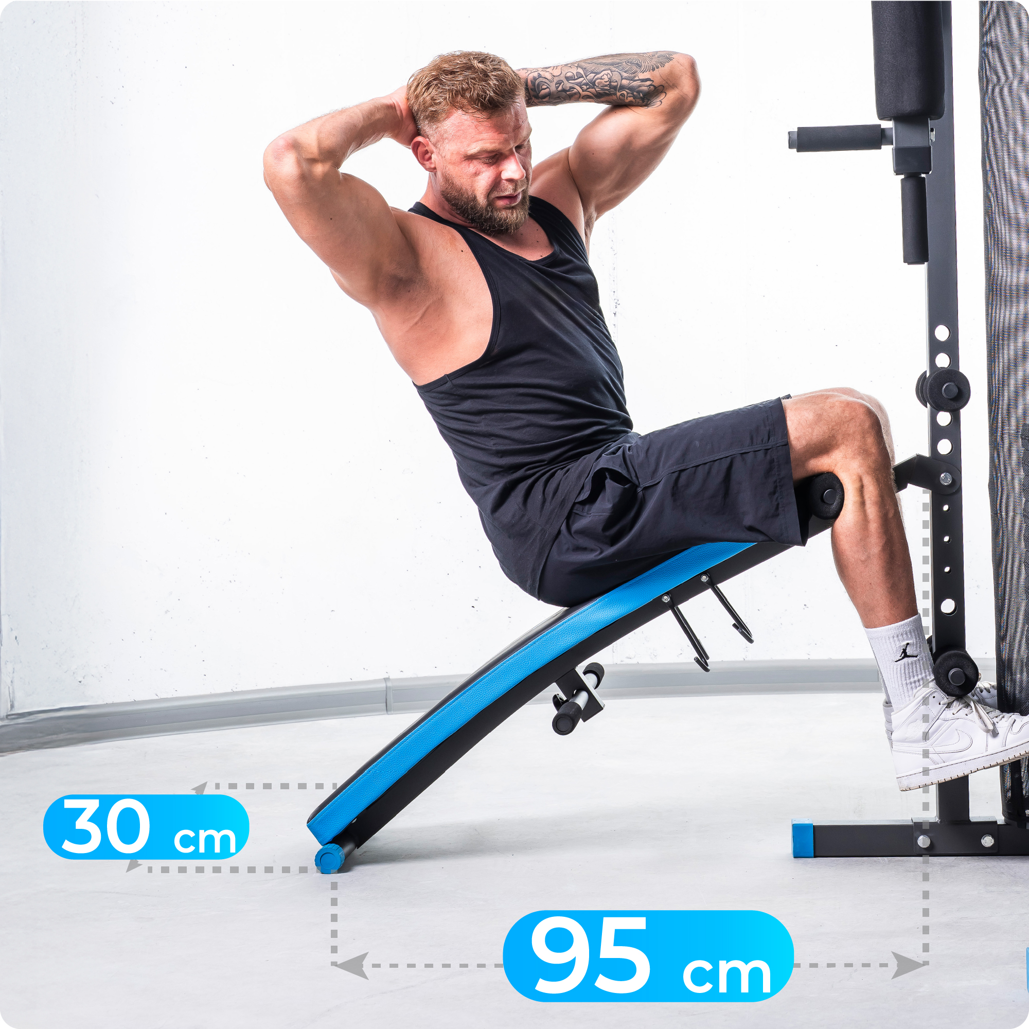 Mężczyzna robi brzuszki na niebiesko-czarnej ławce. Nosi czarne szorty i top. Za nim jest maszyna do ćwiczeń.