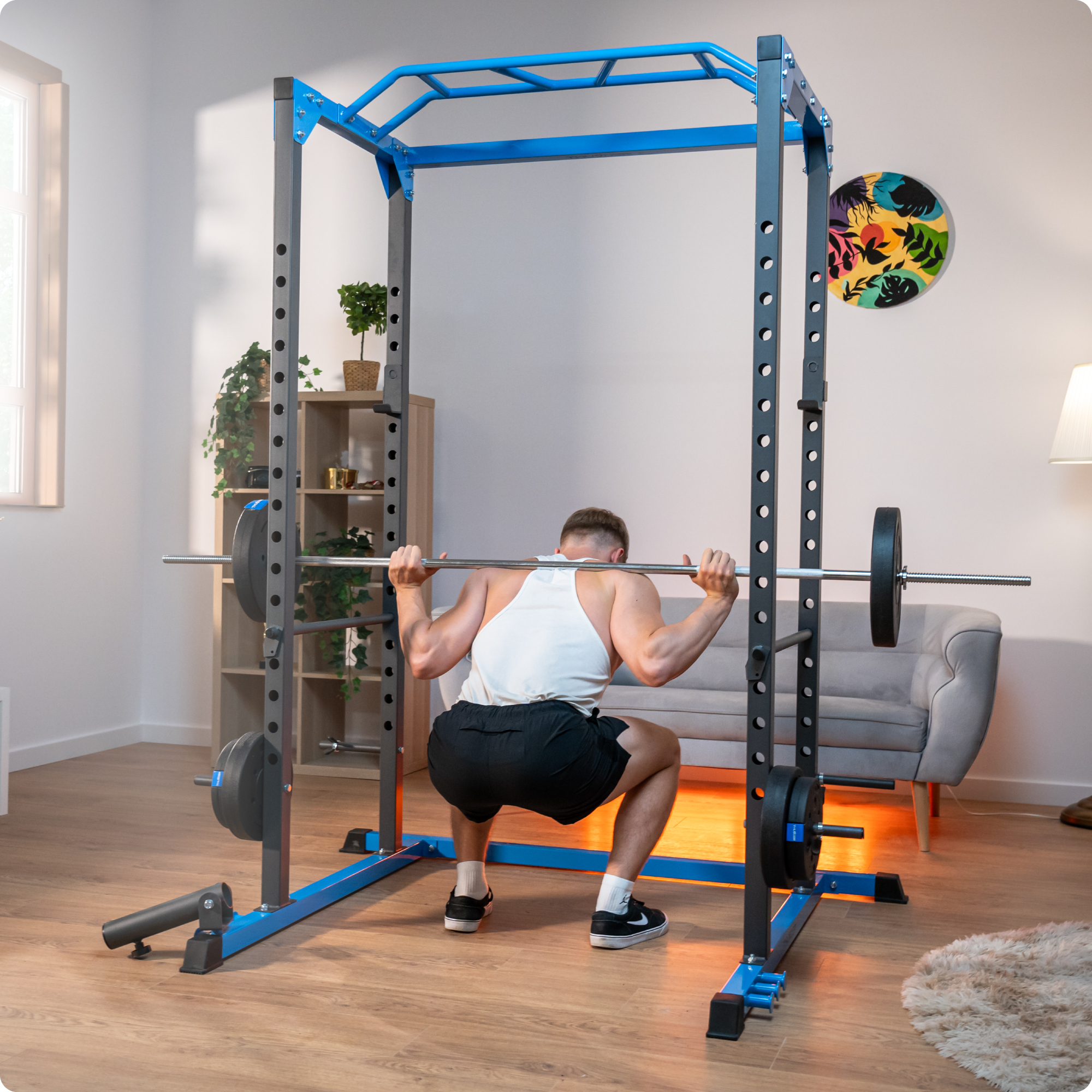 Mężczyzna robi przysiady ze sztangą w domowej siłowni. Widoczne są również stojak do przysiadów, obciążniki, sofa i dekoracje.