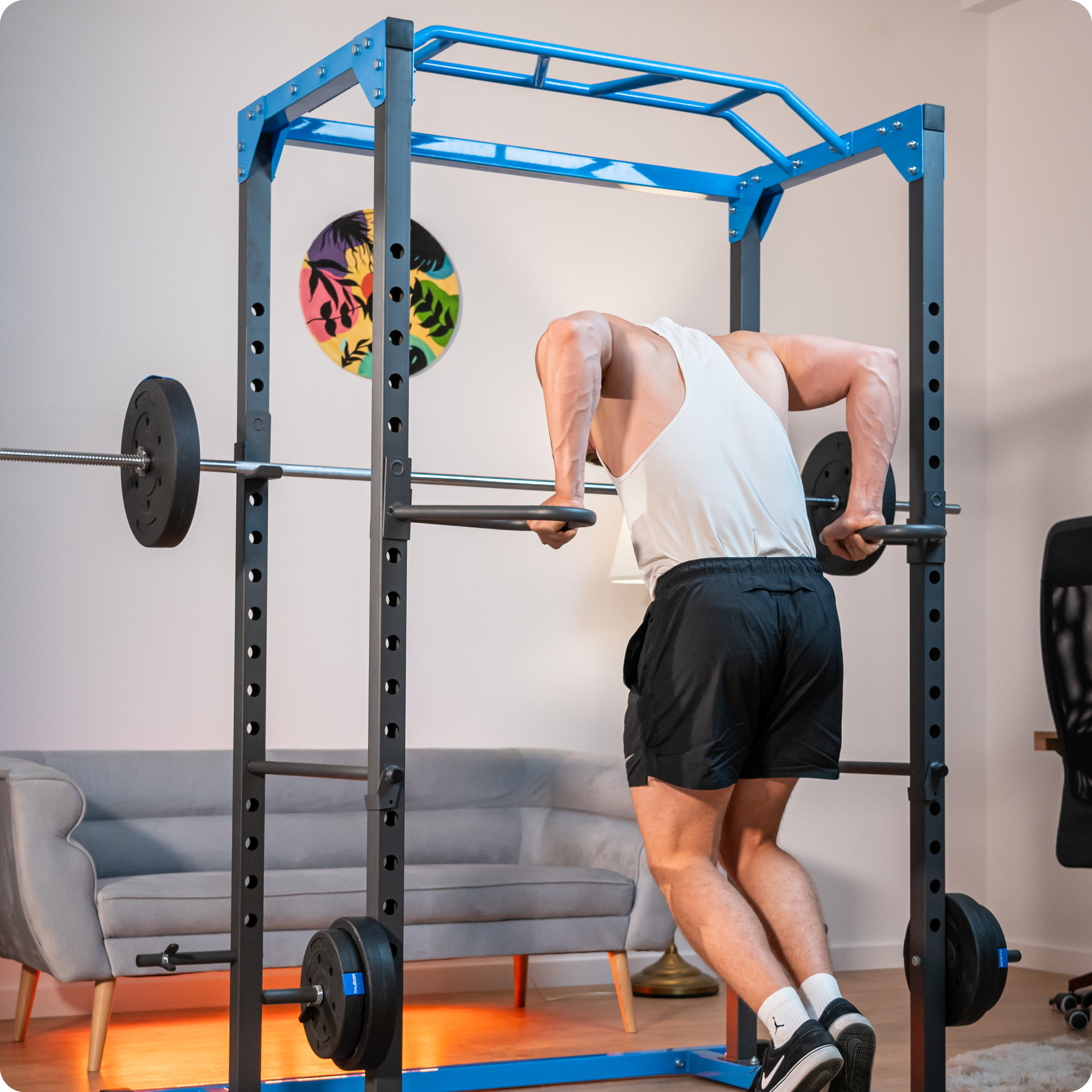 Mężczyzna w białym podkoszulku robi pompki na niebiesko-czarnym stojaku na ciężary. Widoczna jest sztanga z ciężarami.