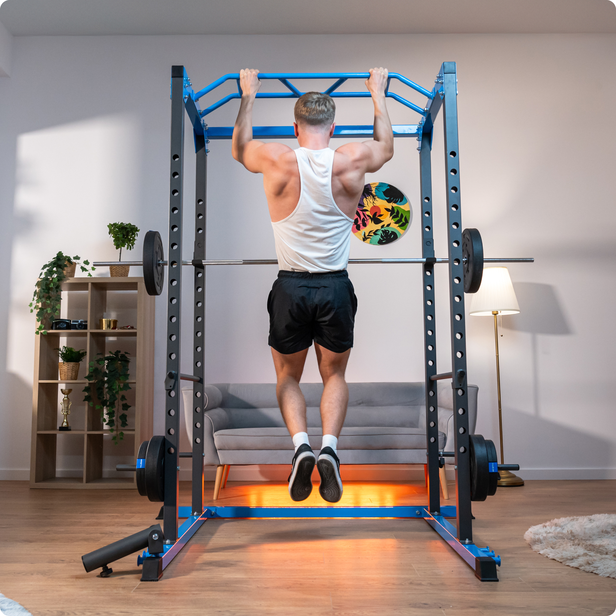 Mężczyzna robi podciągania w domowej siłowni. Ma na sobie biały podkoszulek i czarne szorty. Obciążniki na stojaku.