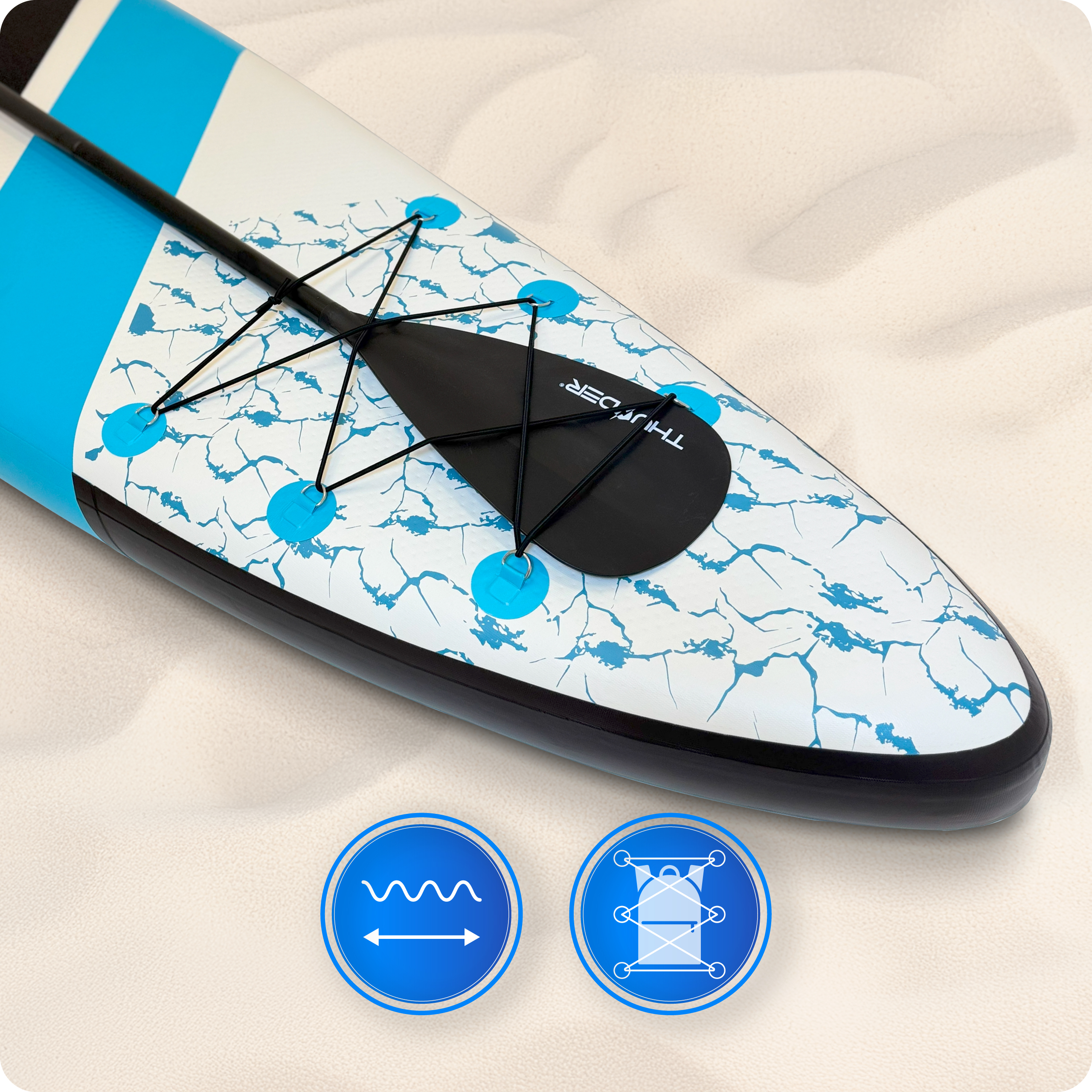 Deska do wiosłowania na piasku. Deska jest biała, niebieska i czarna. Wiosło jest czarne. Dwa niebieskie koła z ikonami na pierwszym planie.