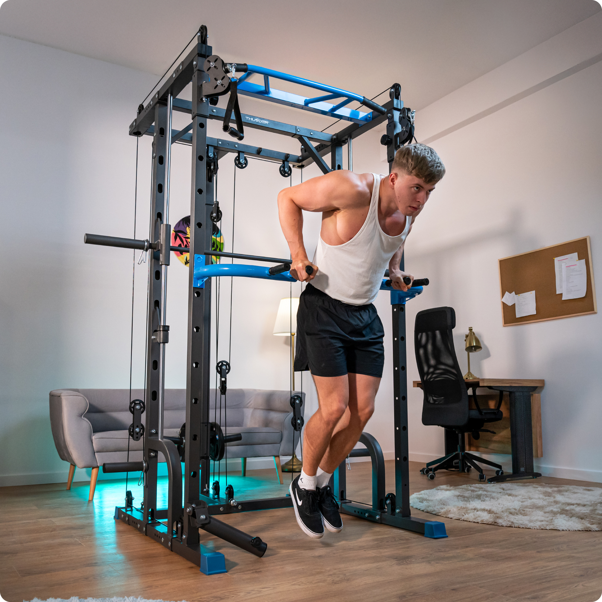 Mężczyzna robi pompki na maszynie do ćwiczeń. Ma na sobie biały podkoszulek i czarne szorty. W tle pokój z meblami.