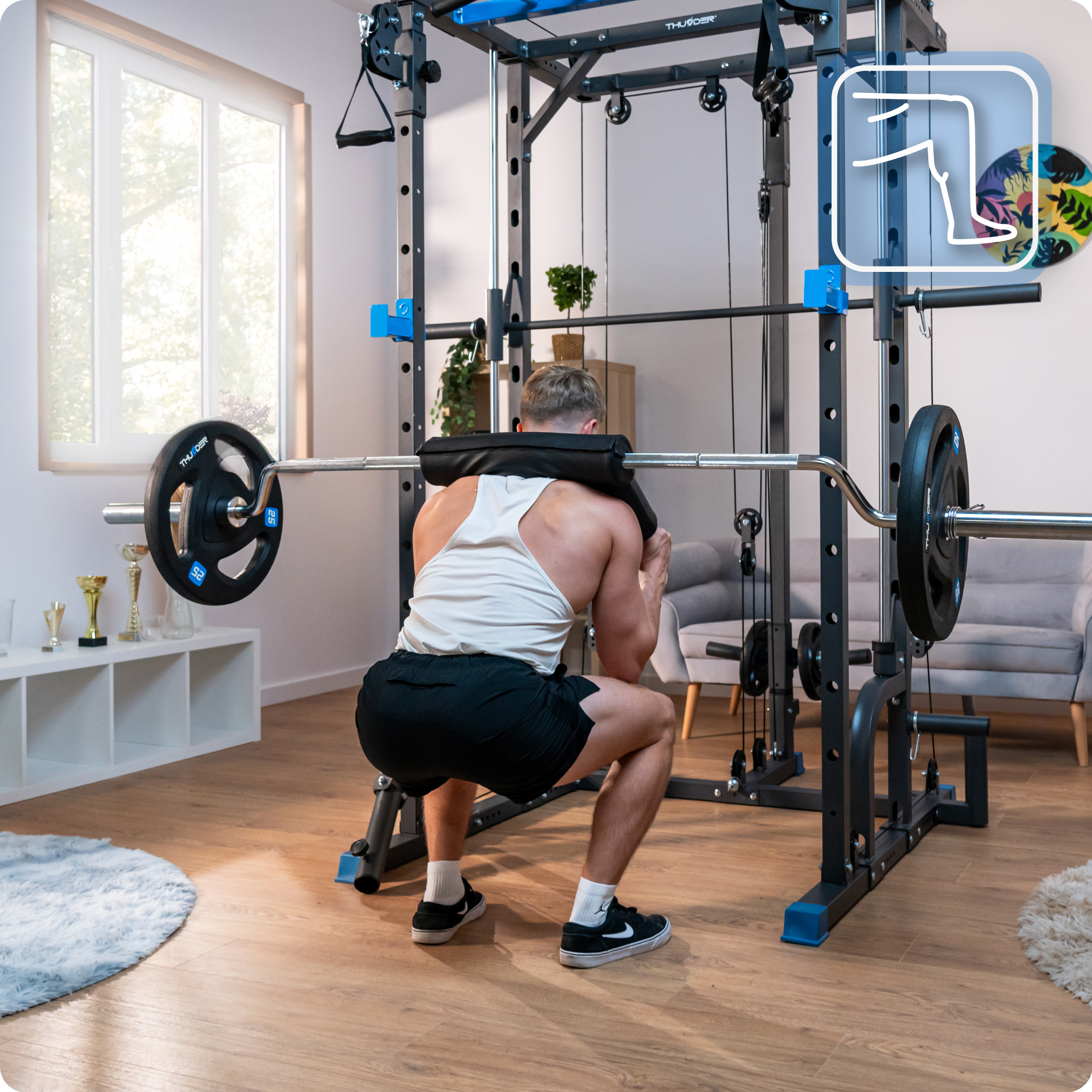 Mężczyzna kuca z sztangą na ramionach. Za nim maszyna do ćwiczeń. Pokój ma okno i drewnianą podłogę.