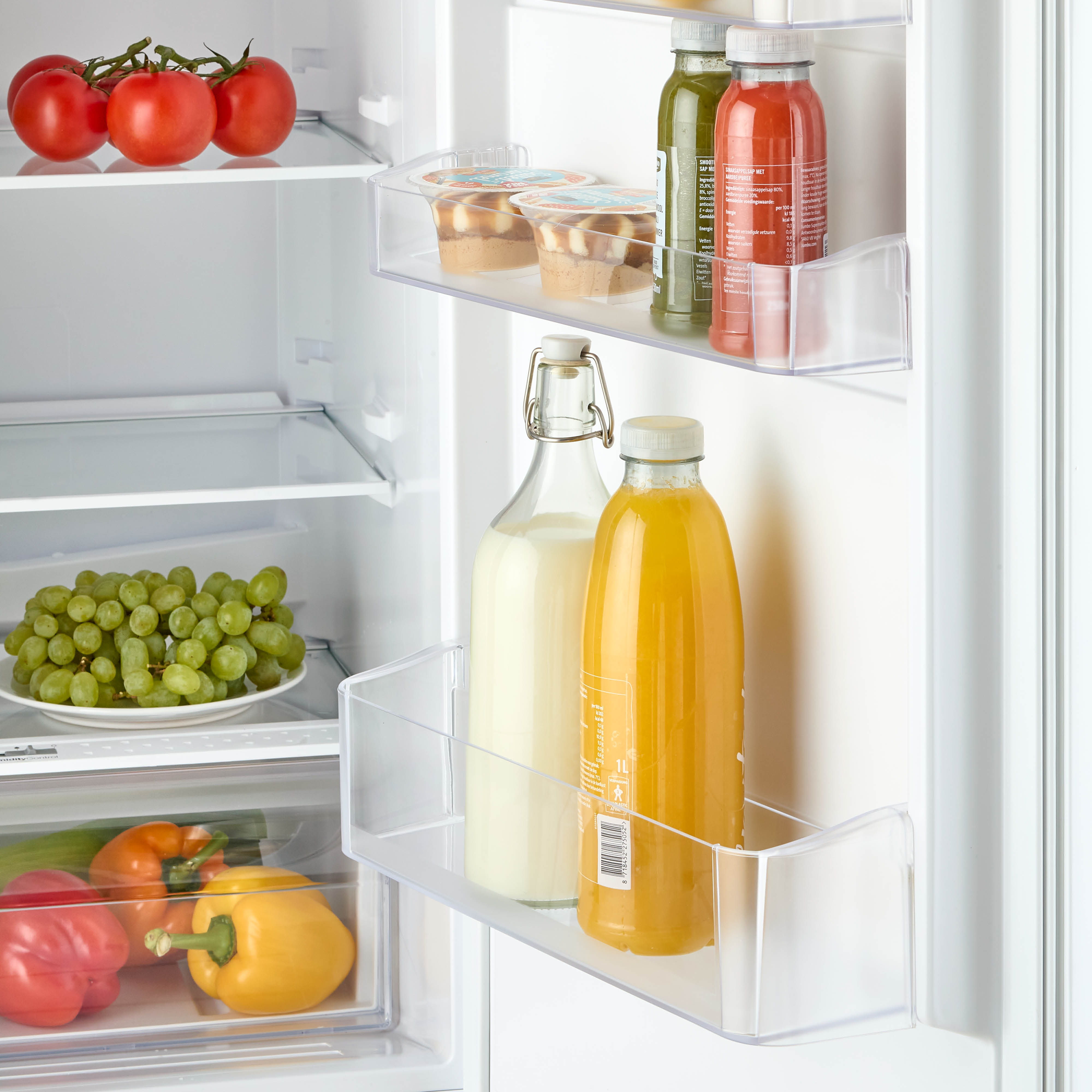 In een koelkast: tomaten, druiven, paprika's, yoghurt, sapflessen en melk. De koelkast is schoon en goed georganiseerd.
