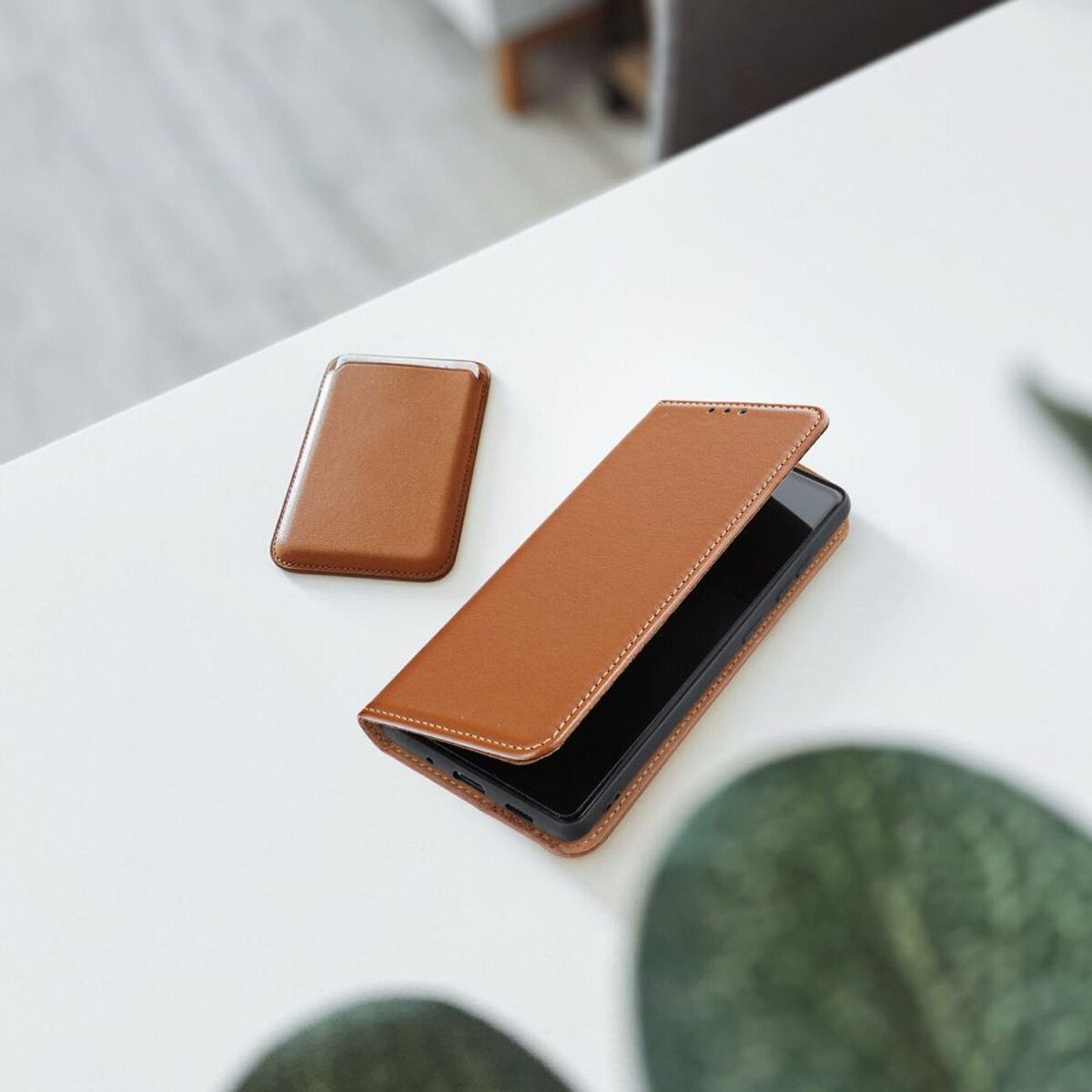 Eine braune Leder-Handyhülle mit einem Telefon darin und einem Kartenhalter auf einem weißen Tisch. Grüne Blätter in der Ecke.