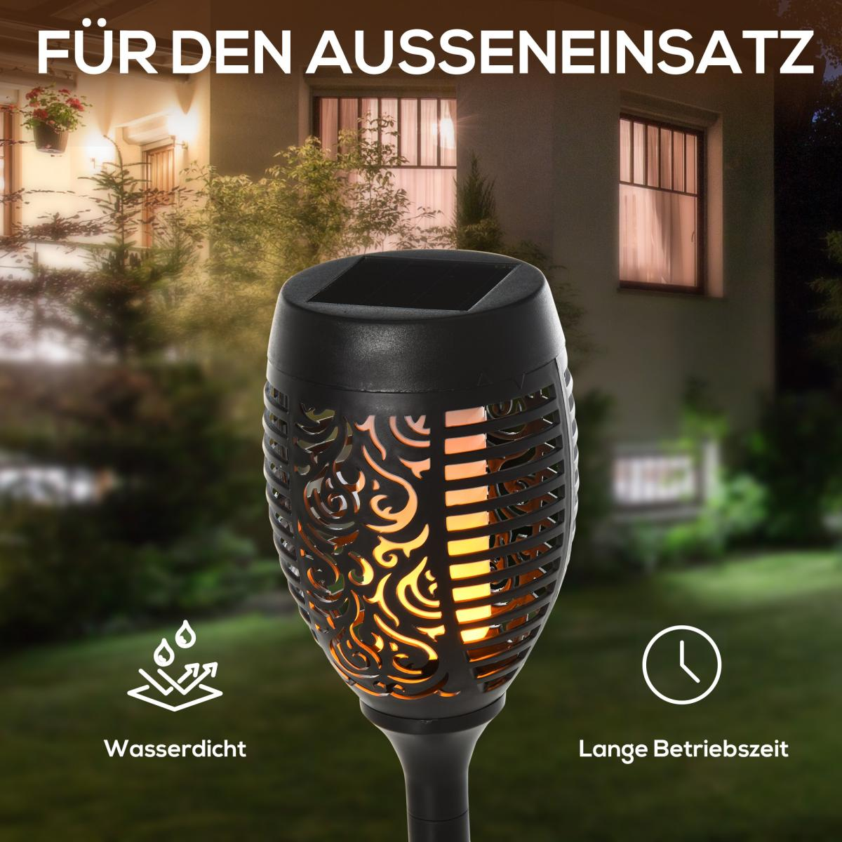 Eine schwarze Solarfackel mit dekorativen Ausschnitten im Vordergrund. Der Hintergrund zeigt ein Haus und Garten bei Nacht.