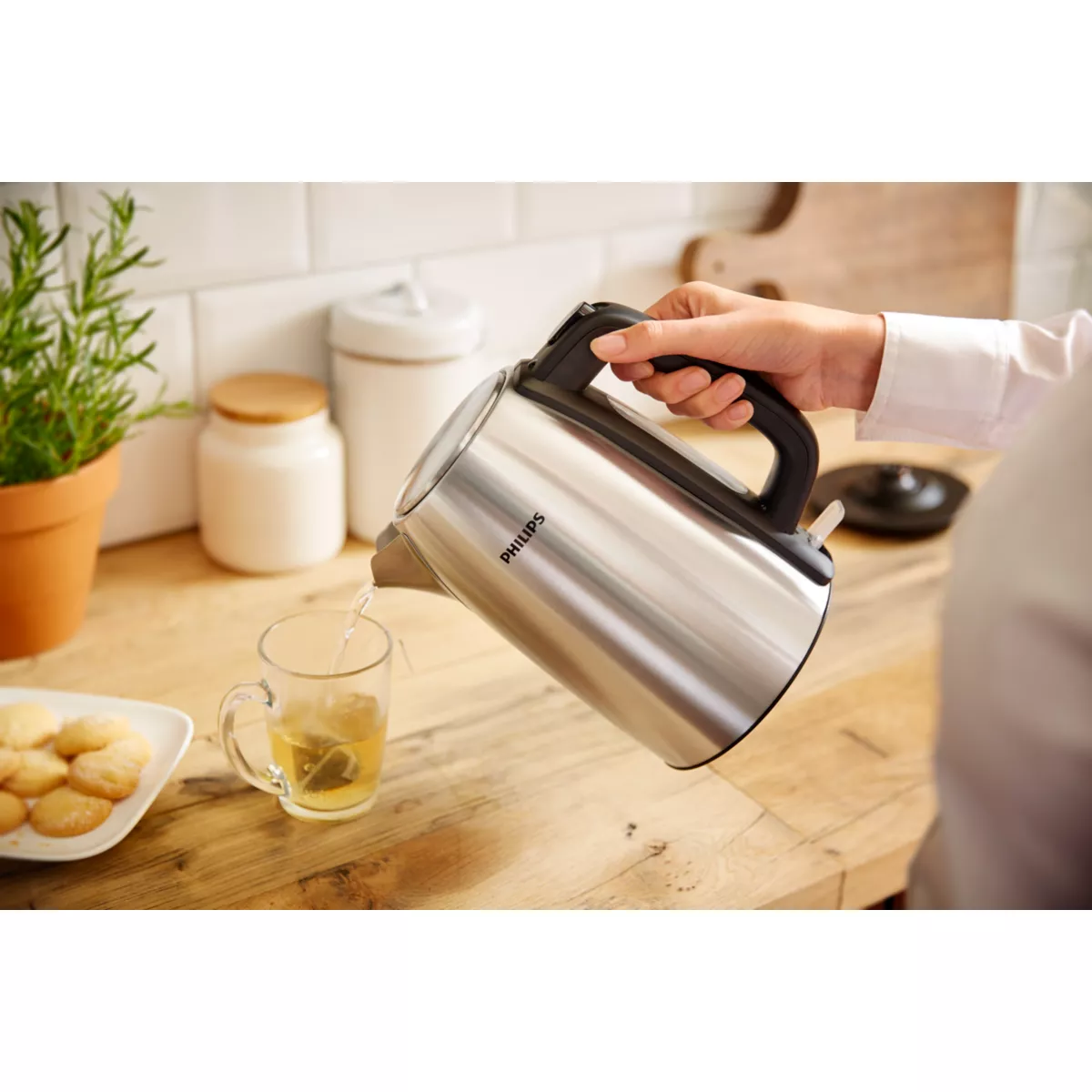 Eine Hand gießt Wasser aus einem Philips-Wasserkocher aus Edelstahl in eine Glas-Tasse mit Teebeutel. Kekse und eine Pflanze sind zu sehen.