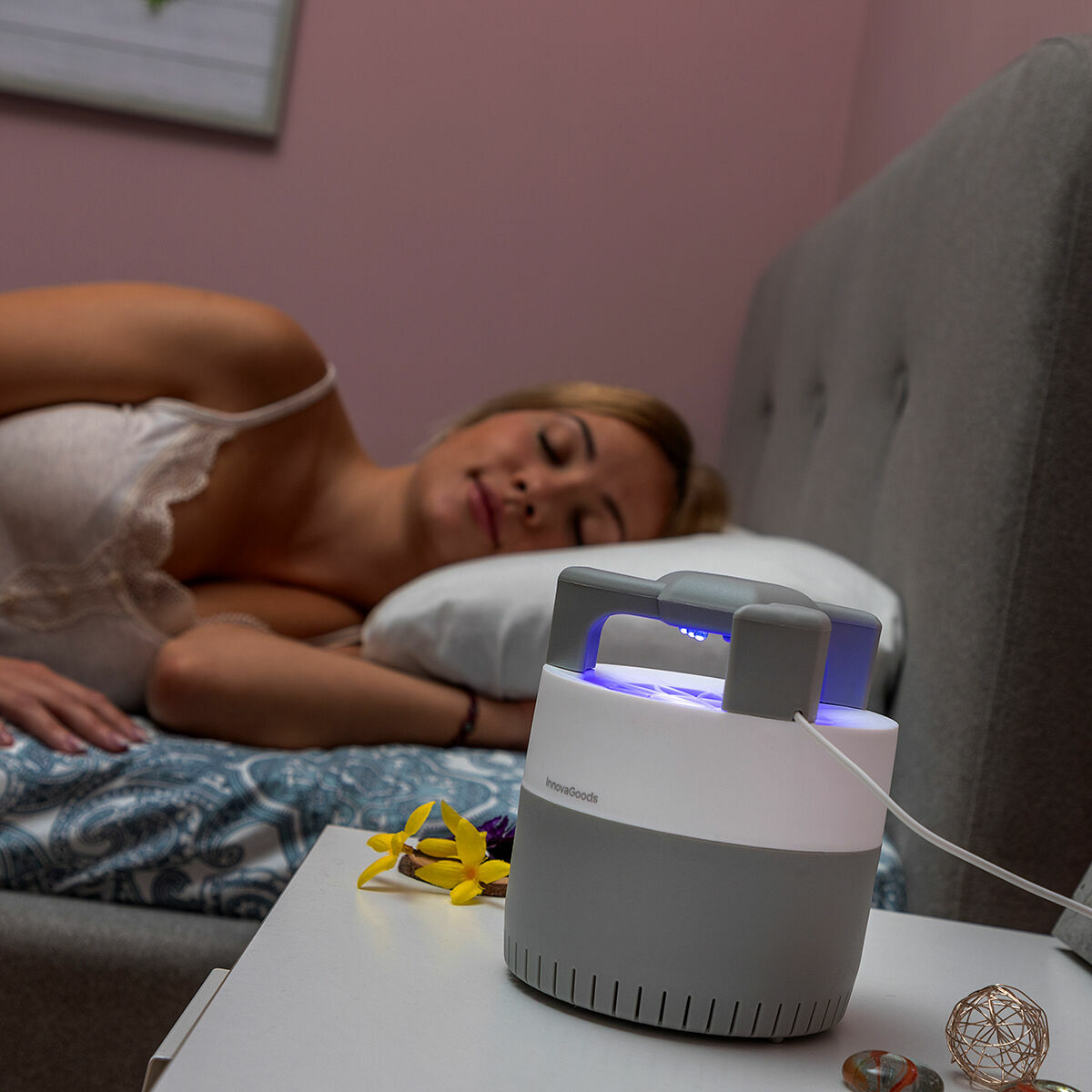 Una donna dorme a letto. Una trappola per zanzare si illumina su un comodino. Fiori gialli sono anche sul comodino.
