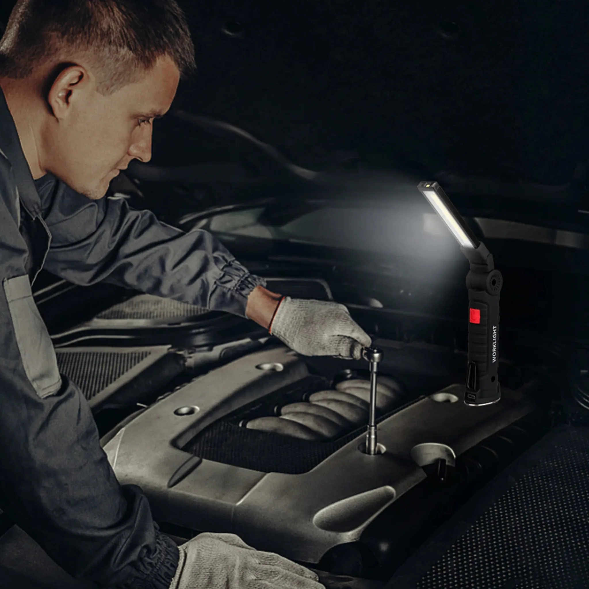 Ein Mechaniker arbeitet mit einem Schraubenschlüssel an einem Automotor. Eine Arbeitsleuchte beleuchtet die Szene. Der Mechaniker trägt Handschuhe.