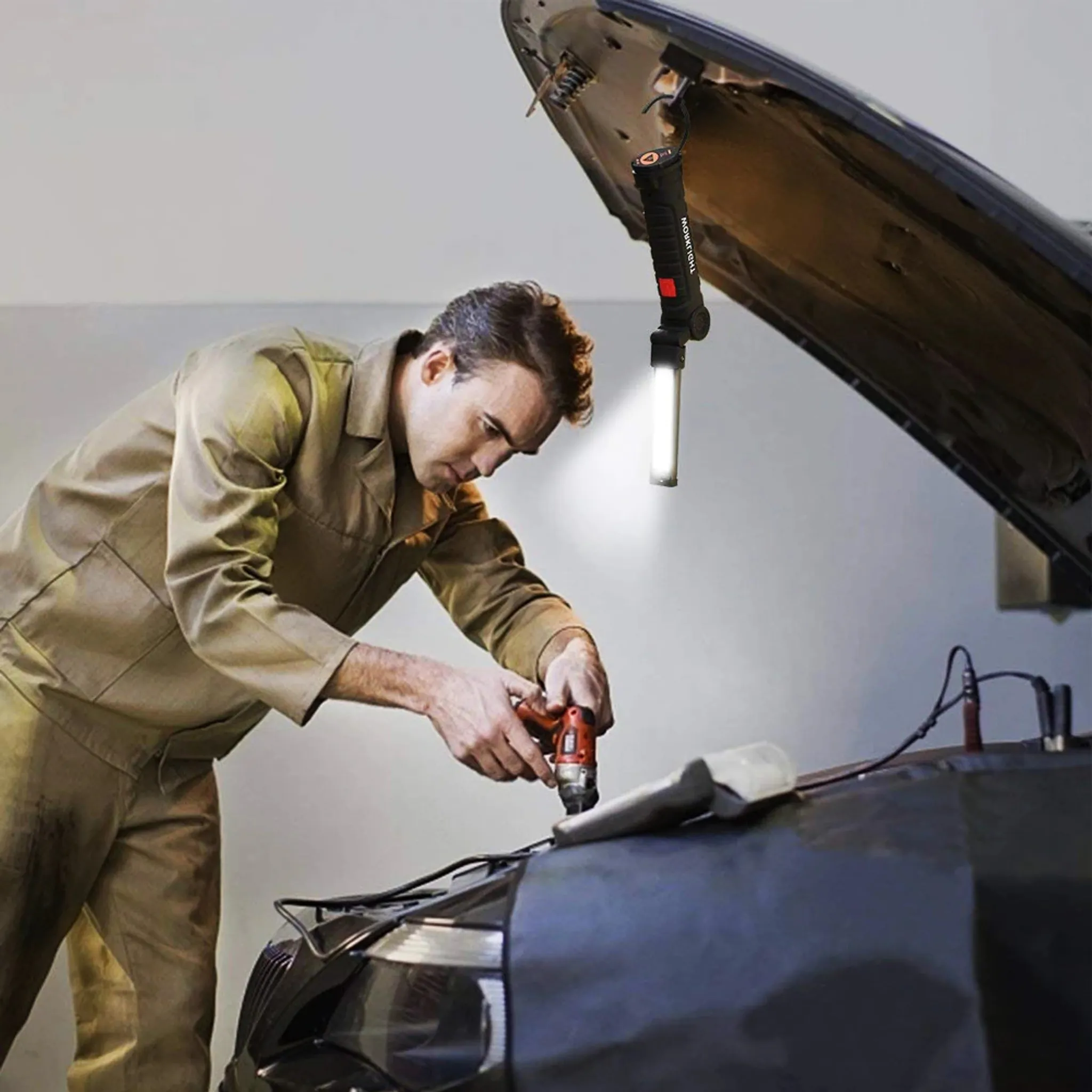 Mechaniker in beiger Uniform repariert einen Automotor unter der Motorhaube, beleuchtet von einer Hängelampe.