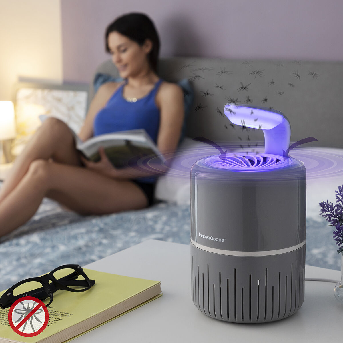 Eine Frau liest im Bett in der Nähe einer Mückenfalle. Die Falle ist grau mit einem violetten Licht. Auf einem Tisch liegen eine Brille und ein Buch.