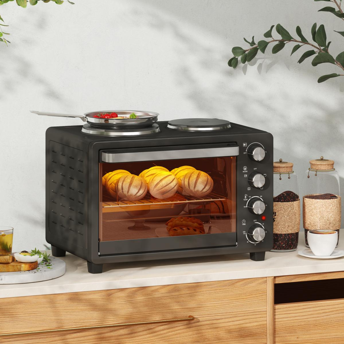 Ein schwarzer Ofen mit Brot darin und eine Pfanne mit Essen oben drauf. Zwei elektrische Kochplatten oben. Gläser und Tasse rechts.