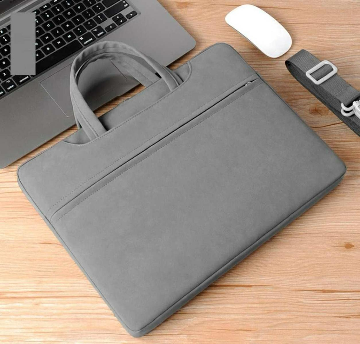 Eine graue Laptoptasche mit Griff, Reißverschluss und Schulterriemen. Ein Laptop, eine weiße Maus, liegen auf Holz.
