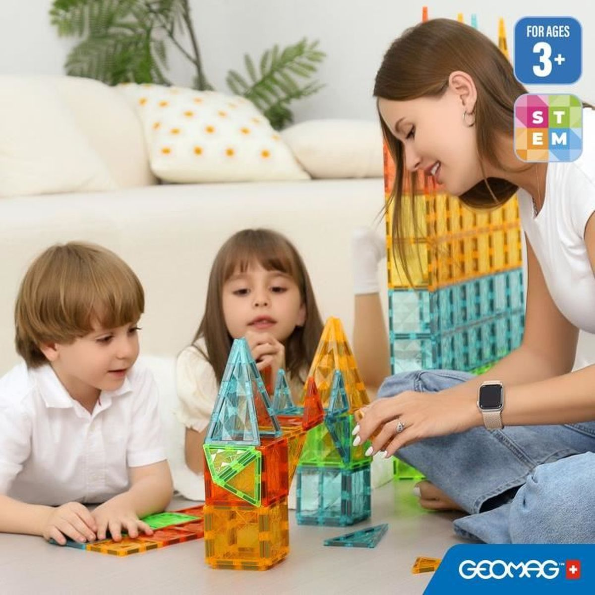 Eine Frau und zwei Kinder spielen mit bunten Magnetbausteinen. Sie bauen einen Turm. Der Hintergrund zeigt ein Sofa.