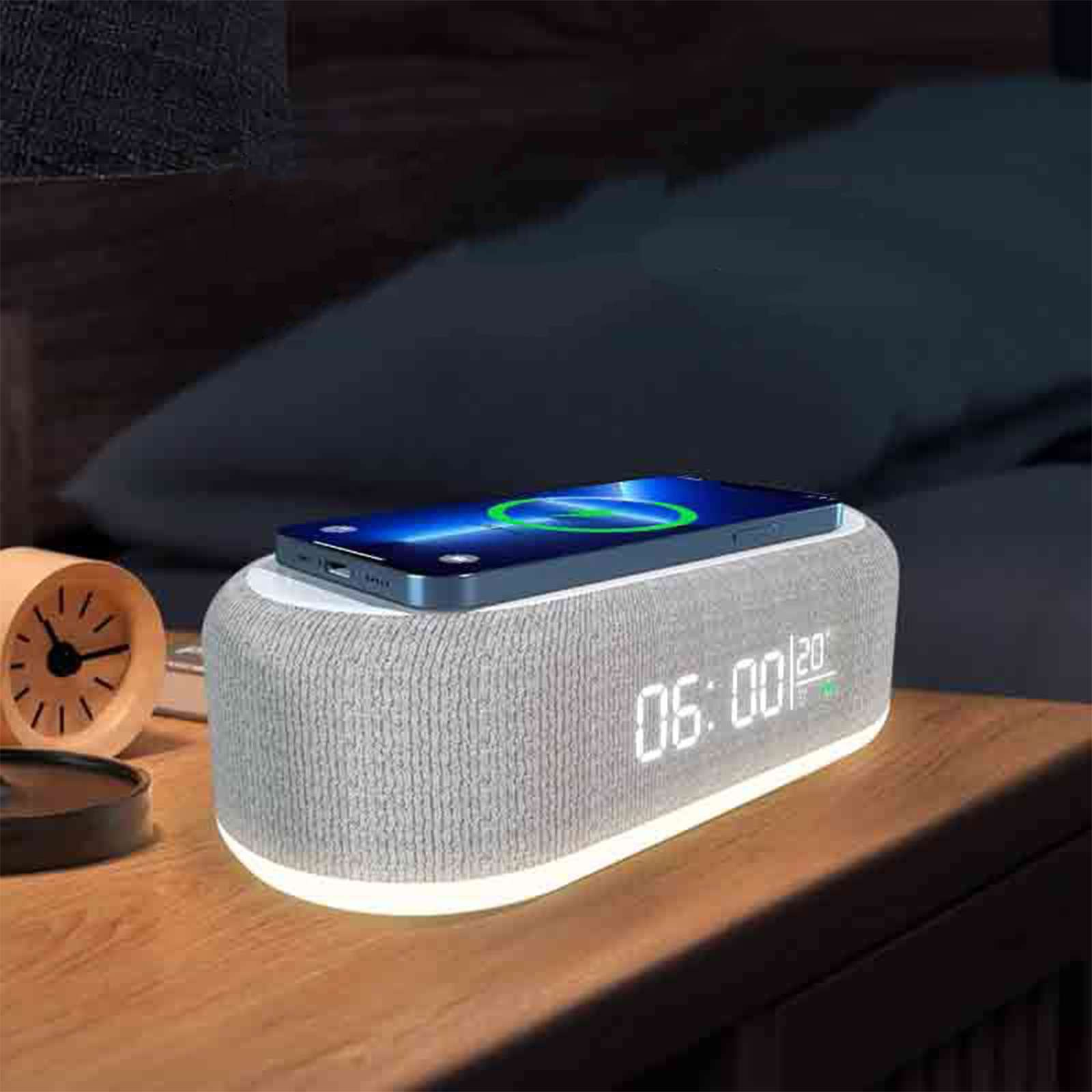 Ein Telefon lädt auf einem Nachttisch. Eine Digitaluhr zeigt 06:00 an. Ein Wecker befindet sich links. Bett im Hintergrund.