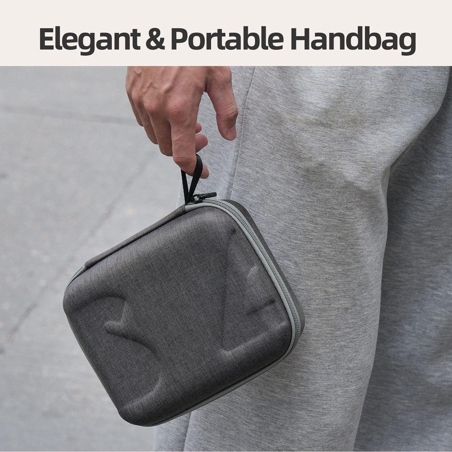 Una mano tiene in mano una borsa grigia con un cinturino nero. La persona indossa pantaloni della tuta grigi. Lo sfondo è una superficie grigia.