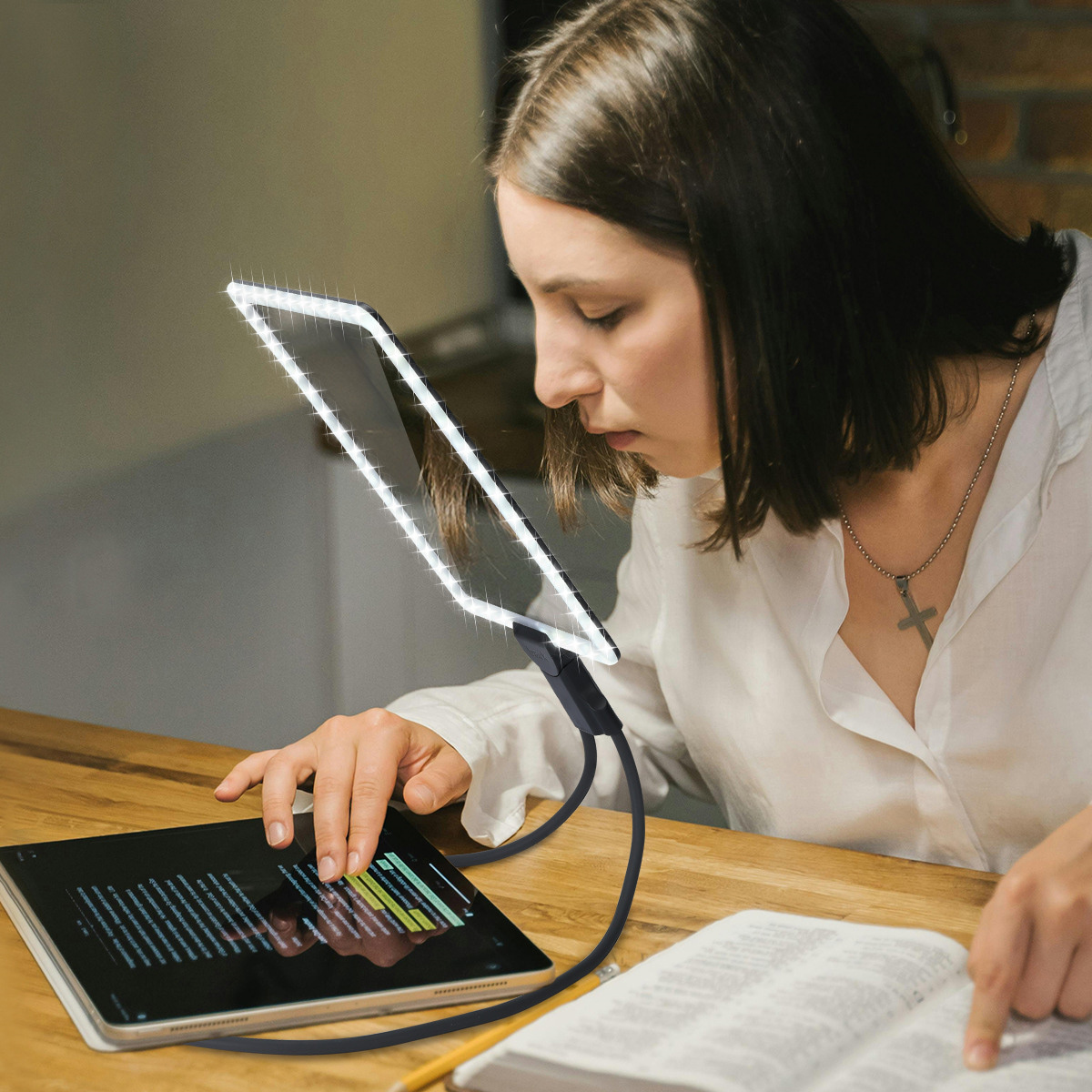Eine Frau liest mit einem beleuchteten Tablet. Eine flexible Lampe leuchtet auf das Tablet. Ein Buch und Bleistift liegen auf dem Tisch.