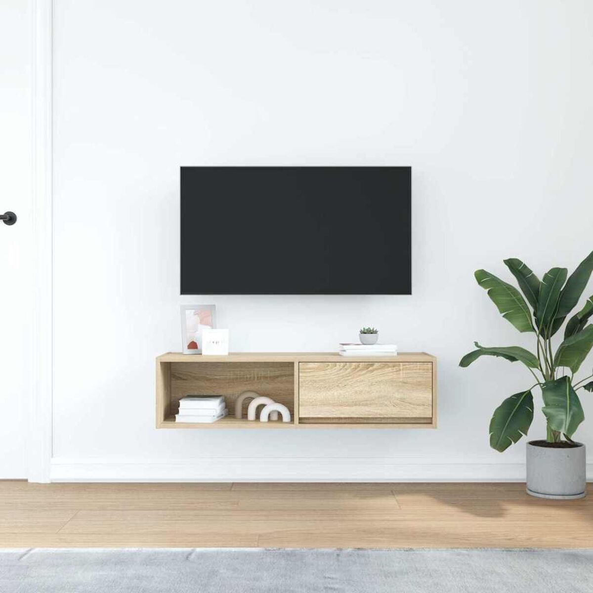 Ein an der Wand montierter Fernseher über einem Holzregal. Rechts steht eine Pflanze. Links ist eine Tür. Der Boden ist aus Holz.
