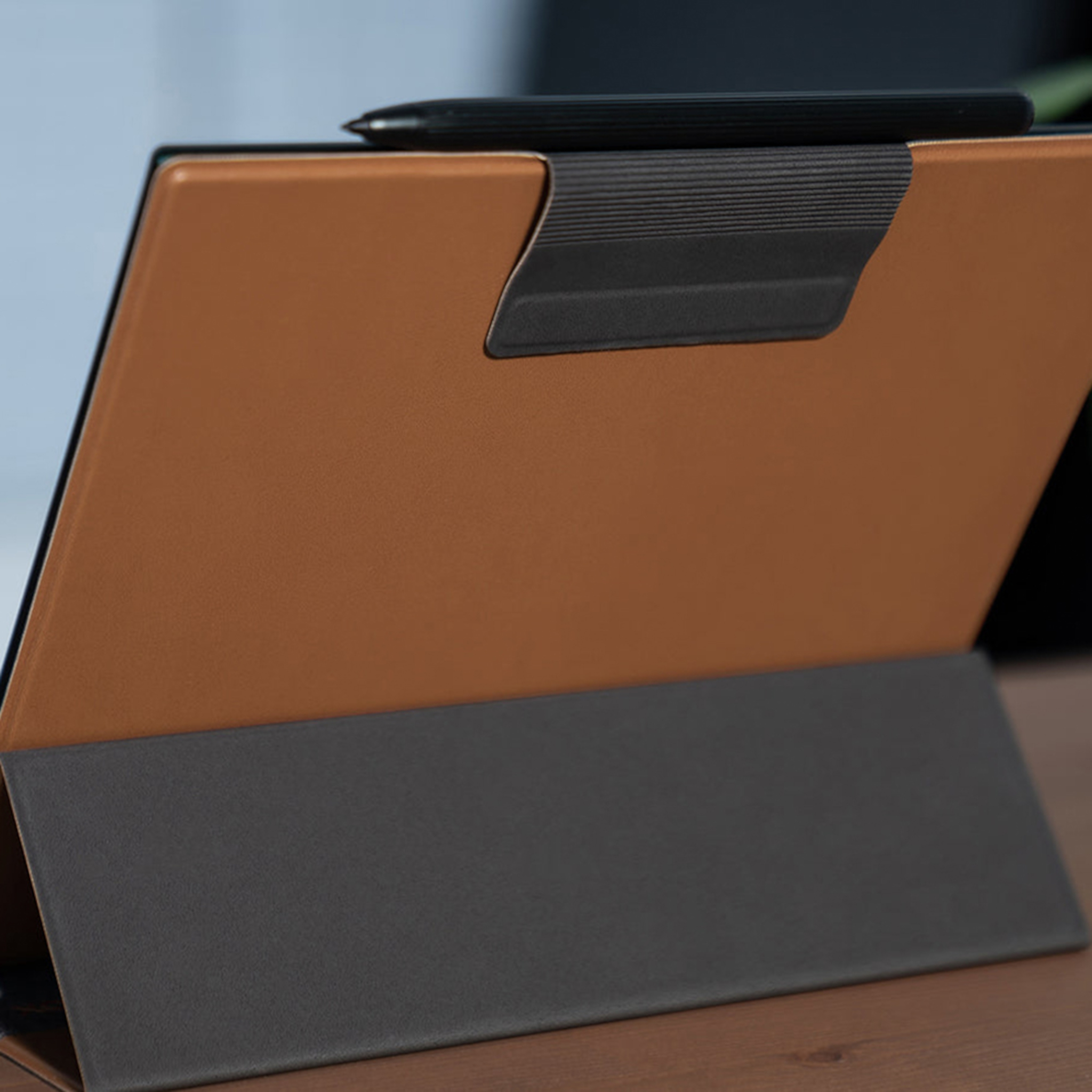 Tablet mit brauner Hülle, schwarzem Ständer und Stift. Es steht auf einem Holztisch. Der Hintergrund ist hellblau.
