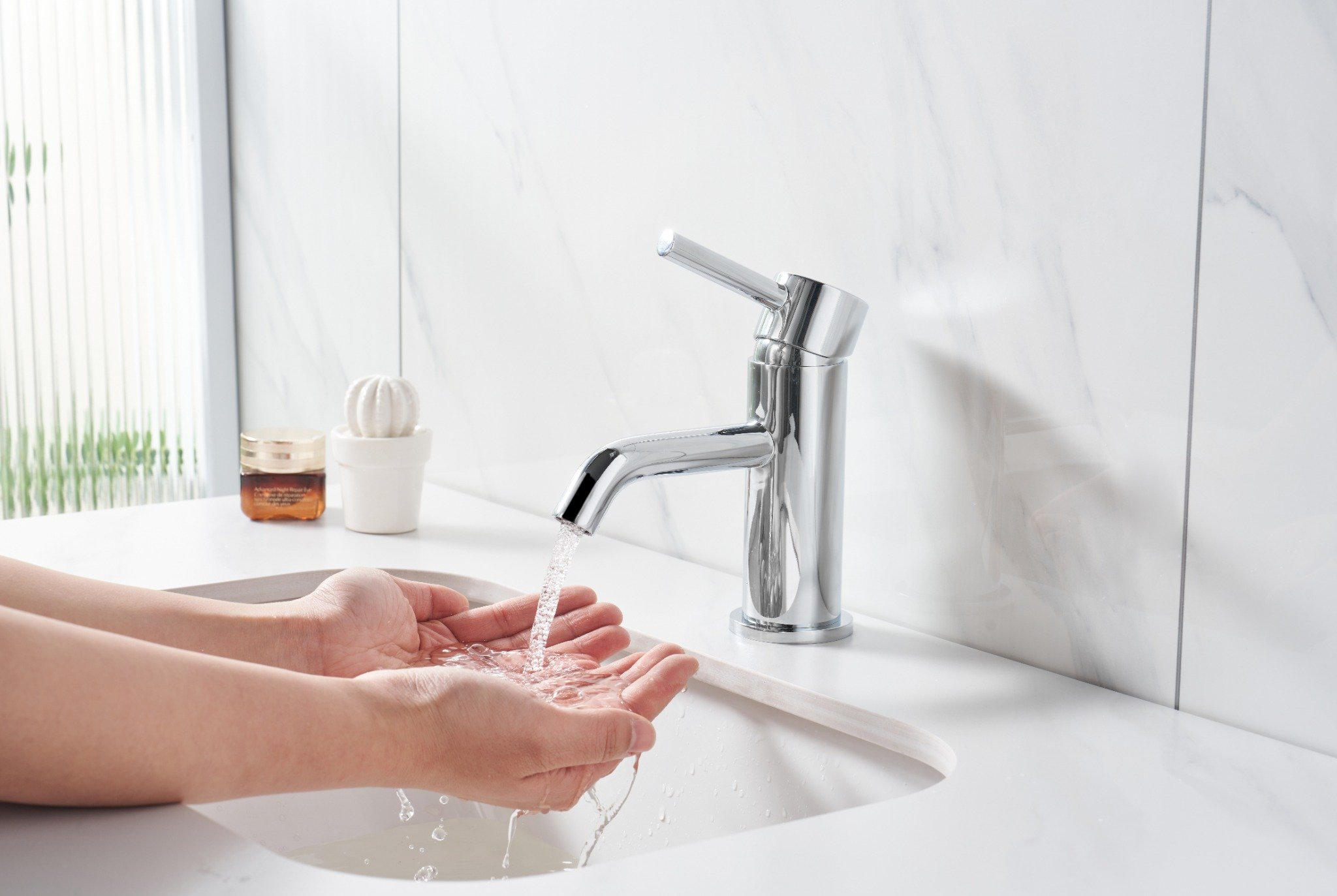Hände unter fließendem Wasser aus einem verchromten Wasserhahn in einem weißen Badezimmer. Ein kleiner Kaktus und ein Glas sind sichtbar.