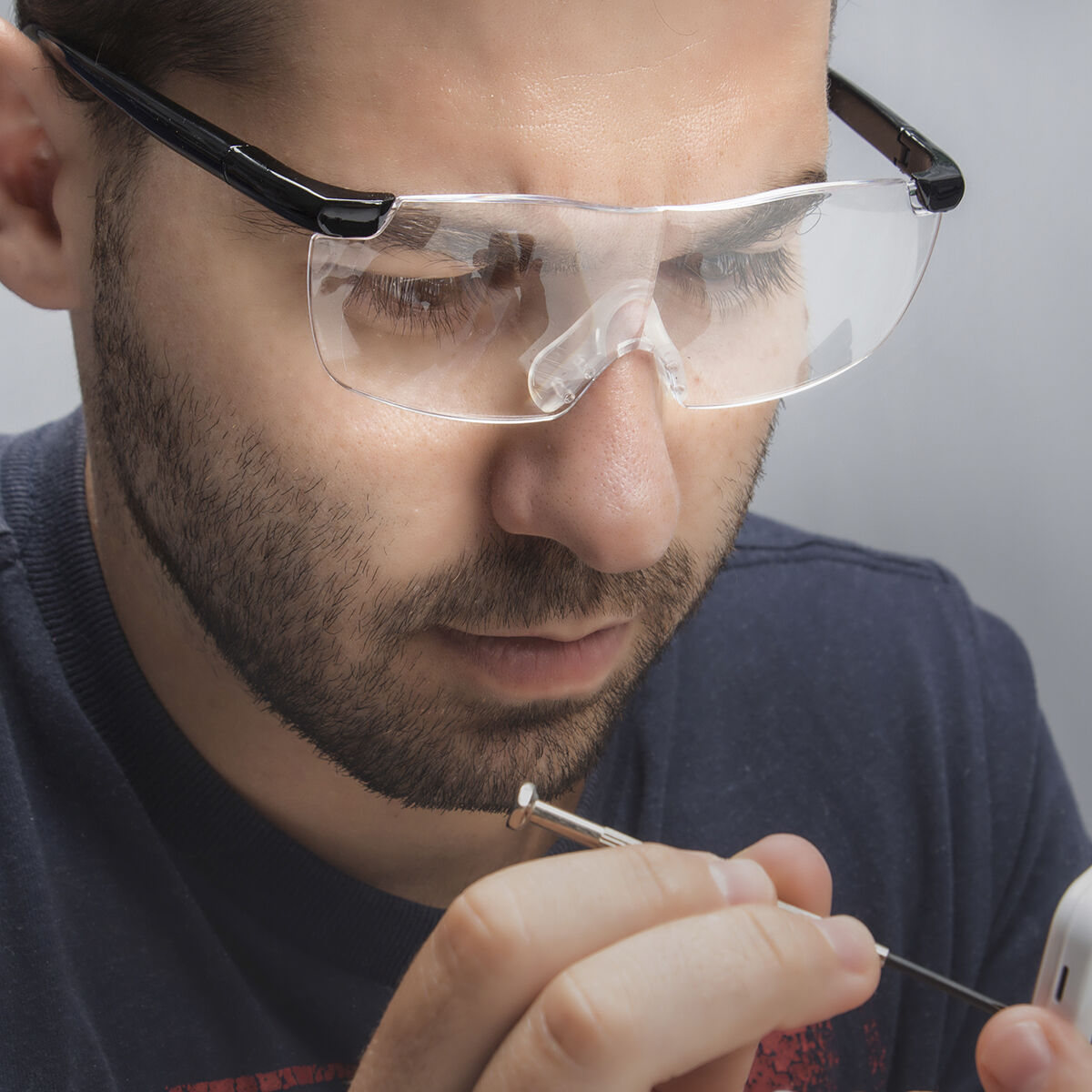 Hombre con gafas de aumento, sosteniendo un destornillador, trabajando en un pequeño dispositivo blanco. Camiseta azul oscuro, fondo gris.