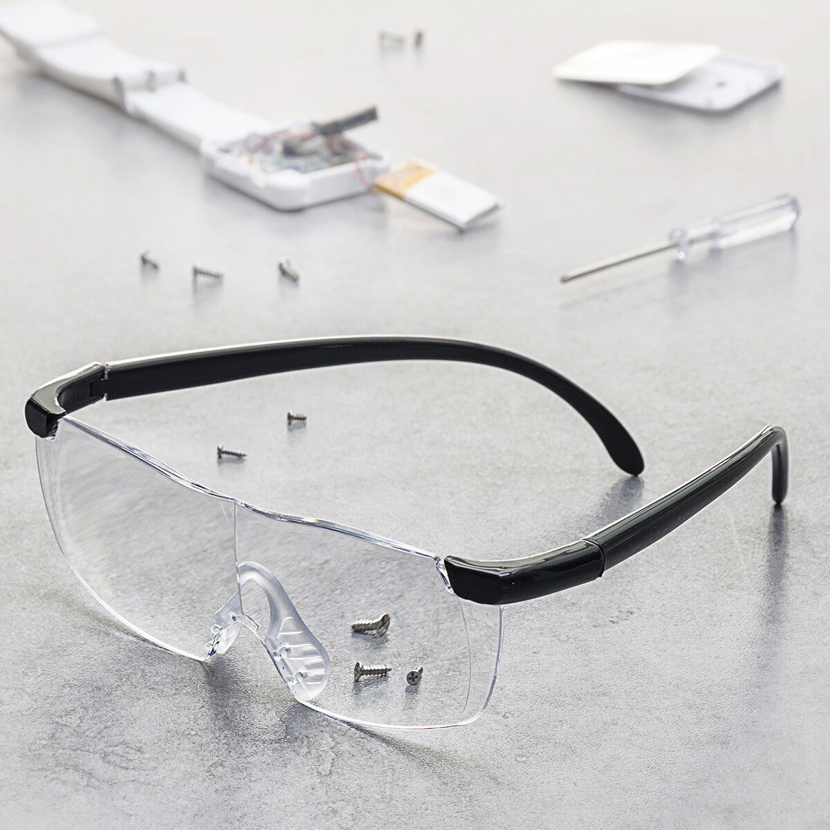 Gafas de seguridad con montura negra y lentes transparentes sobre una superficie gris con tornillos, un dispositivo desmontado y un destornillador.