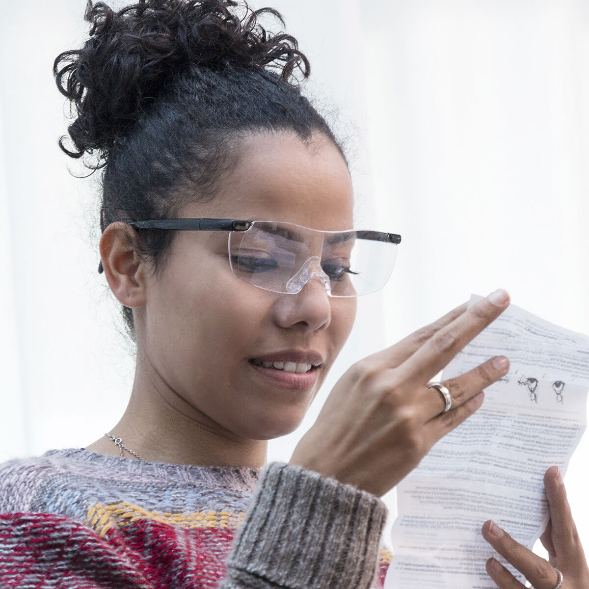 Una mujer con gafas lee un papel. Lleva un suéter y tiene el pelo recogido. El fondo es blanco.