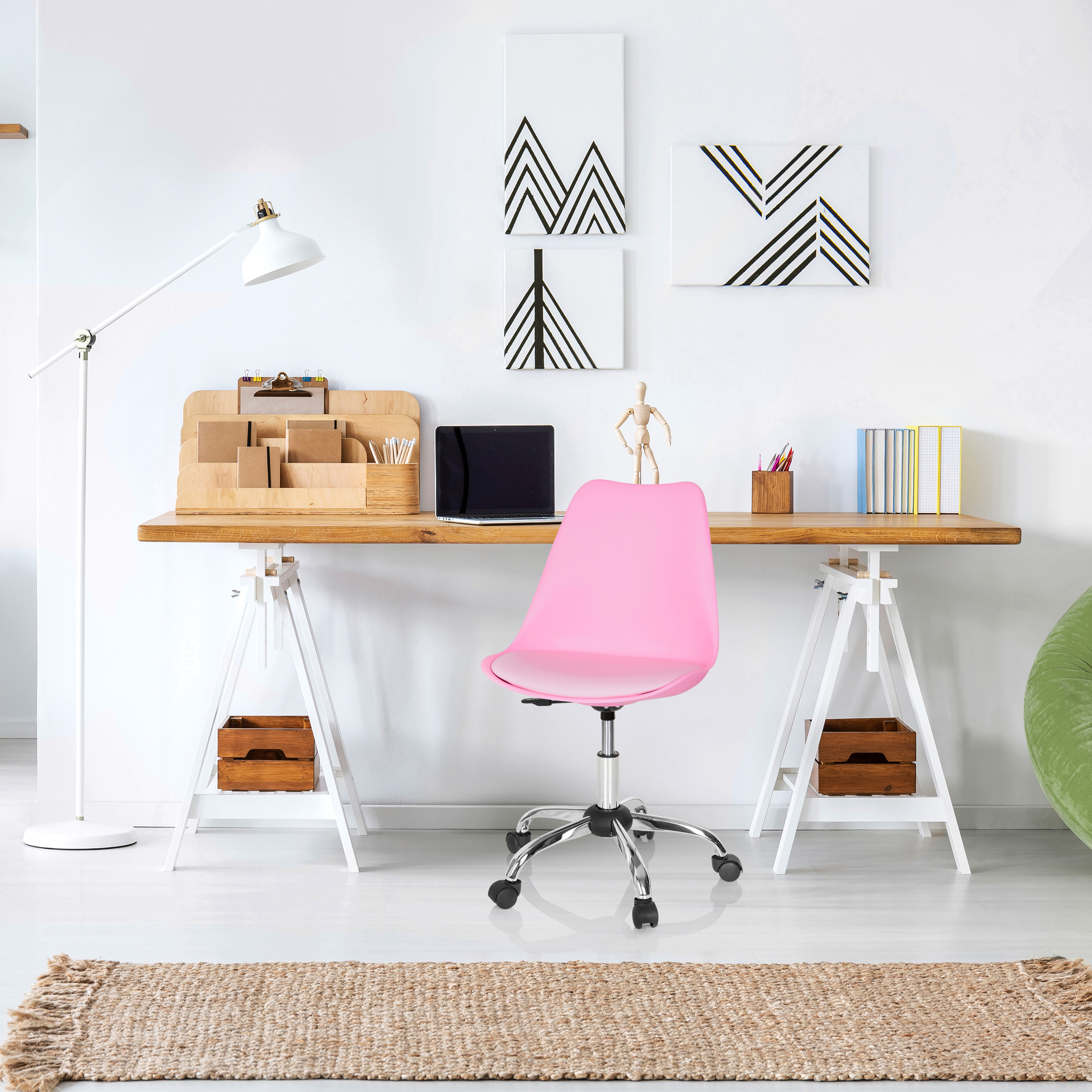 Ein Arbeitsplatz mit Holztisch, rosa Stuhl und weißen Wänden. Es gibt einen Laptop, Kunst und einen Teppich.