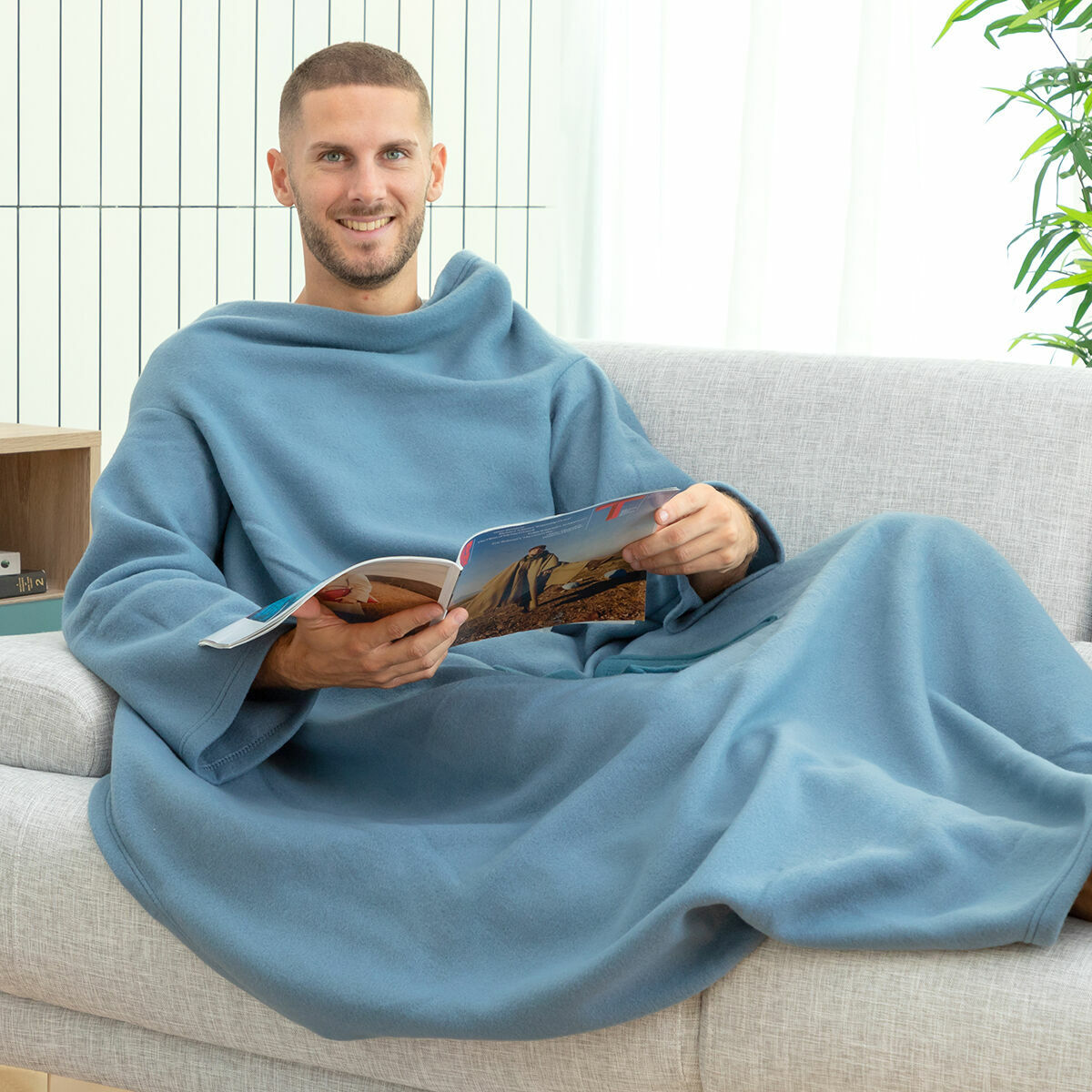 Ein Mann in einer blauen Decke liest eine Zeitschrift auf einem grauen Sofa. Im Hintergrund ist ein Fenster mit weißen Vorhängen.