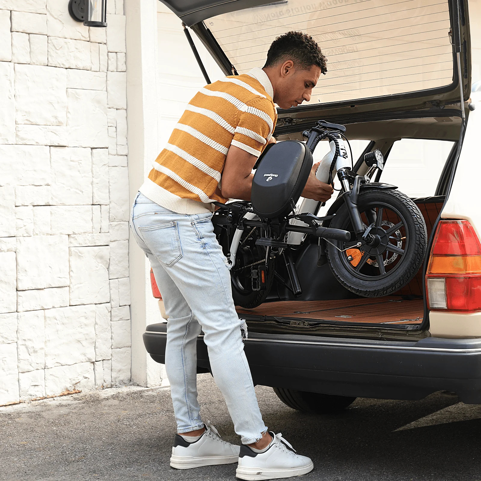 Mężczyzna ładuje złożony rower elektryczny do bagażnika samochodu. Ma na sobie koszulę w paski i dżinsy, stoi obok ceglanego muru.