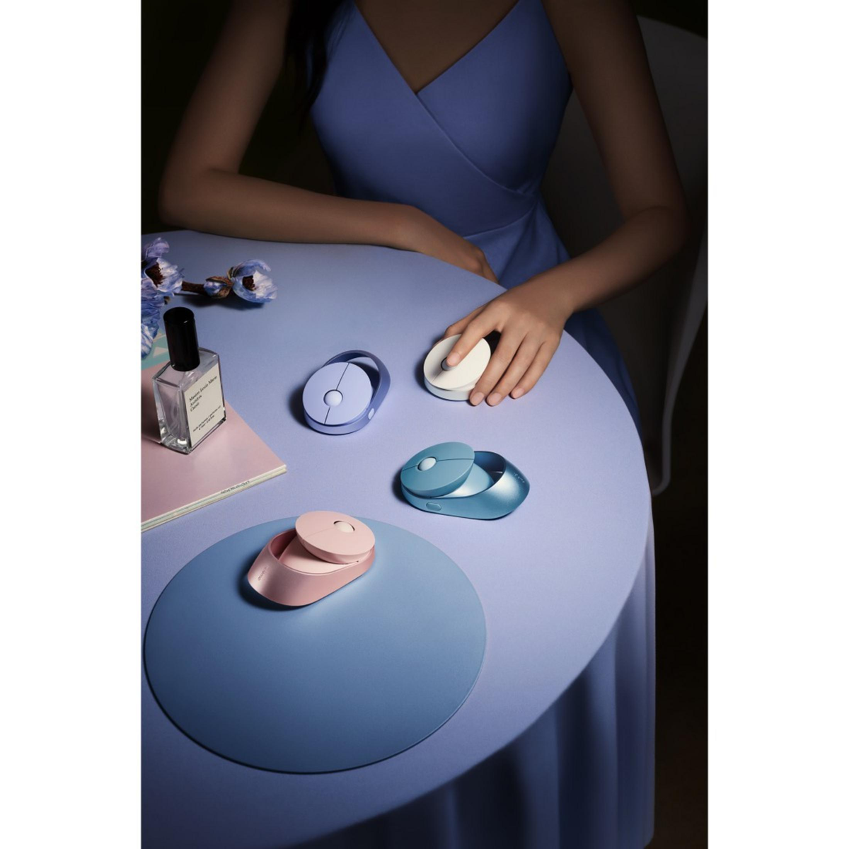 Una mujer en una mesa con varios dispositivos electrónicos pequeños. La mesa es azul y tiene un mantel individual azul y un frasco de perfume.