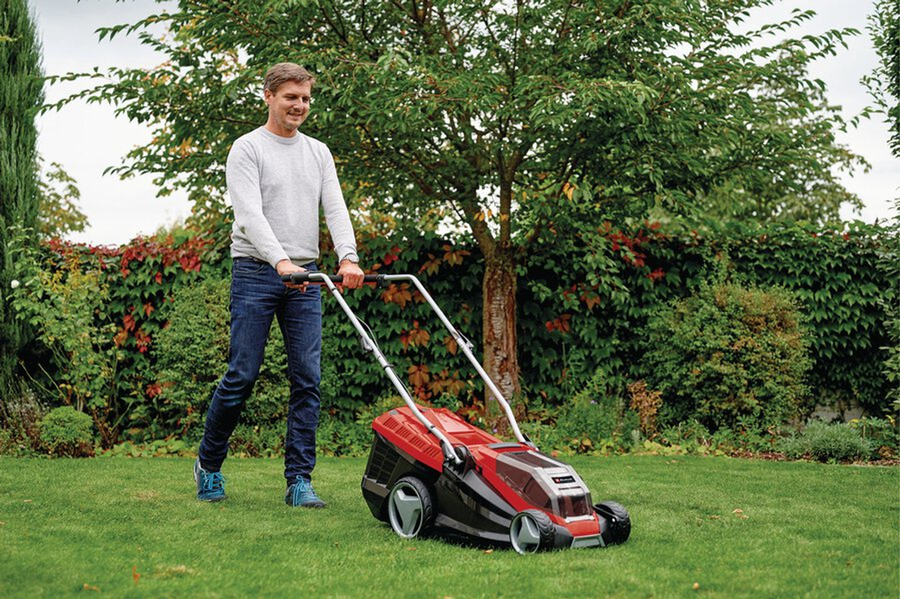 Ein Mann in Jeans und Pullover schiebt einen rot-schwarzen Rasenmäher auf einem grünen Rasen. Bäume und Büsche sind im Hintergrund.