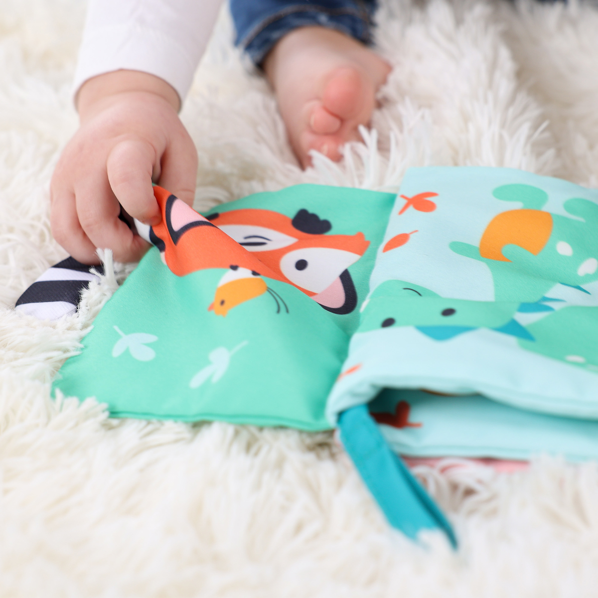 Ręka dziecka sięga po kolorową książeczkę z tkaniny z ilustracjami zwierząt, na puszystej, białej powierzchni. Stopy dziecka widoczne w tle.