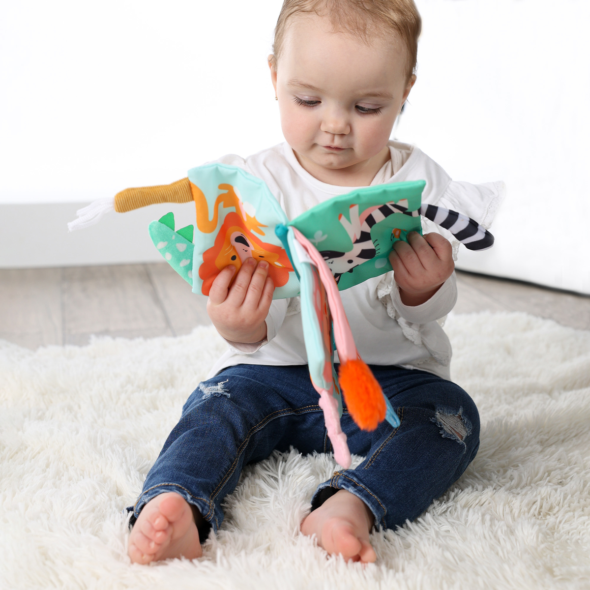 Niemowlę siedzi na puszystym białym dywanie, czytając kolorową miękką książeczkę z ilustracjami zwierząt. Niemowlę ma na sobie dżinsy i białą koszulę.