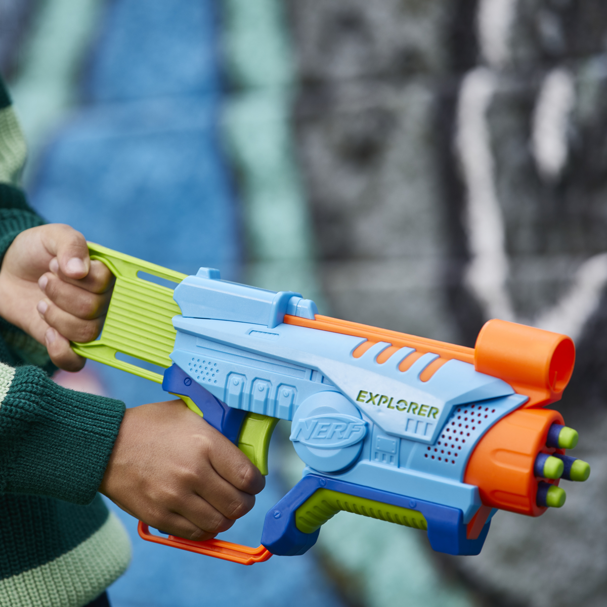 Un bambino tiene in mano una pistola giocattolo Nerf Explorer blu, verde e arancione. Lo sfondo è un muro sfocato con graffiti blu e grigi.