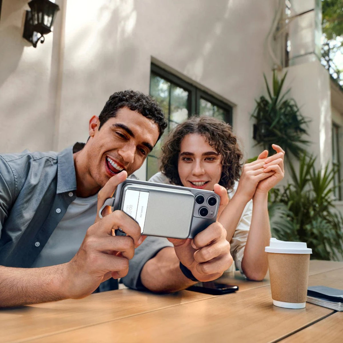 Mężczyzna i kobieta robią sobie selfie telefonem. Siedzą na zewnątrz przy drewnianym stole. W pobliżu stoi filiżanka kawy.