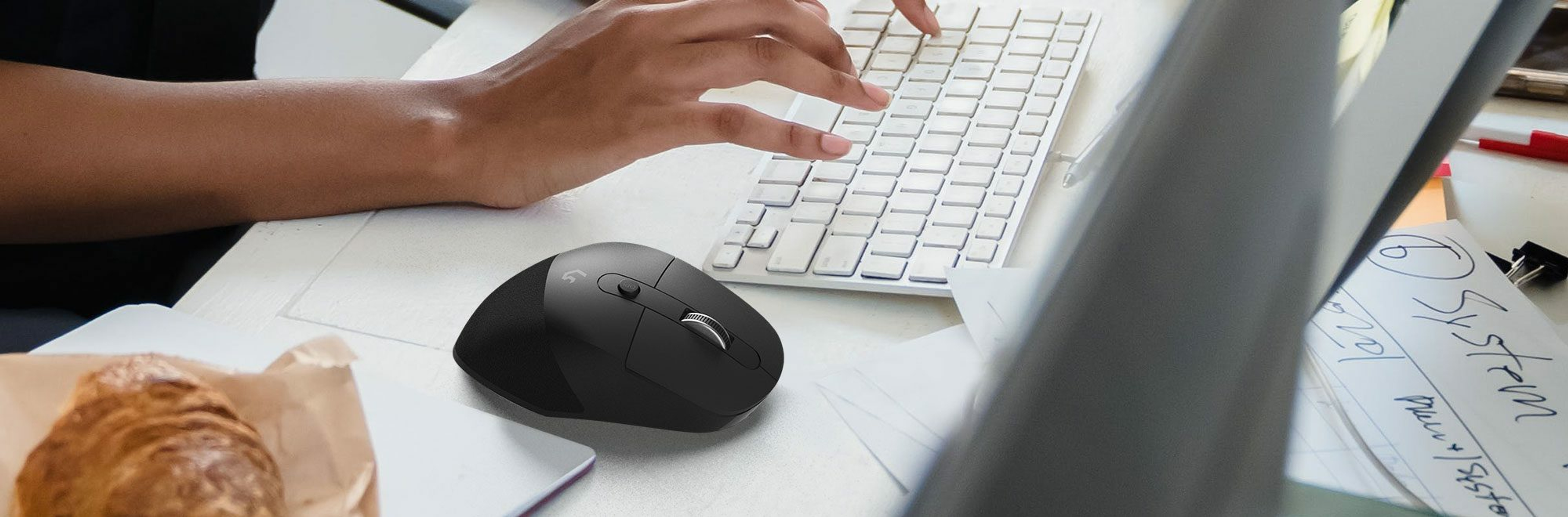 Una mano digita su una tastiera bianca. Un mouse nero è sulla scrivania, accanto a un croissant e dei fogli.