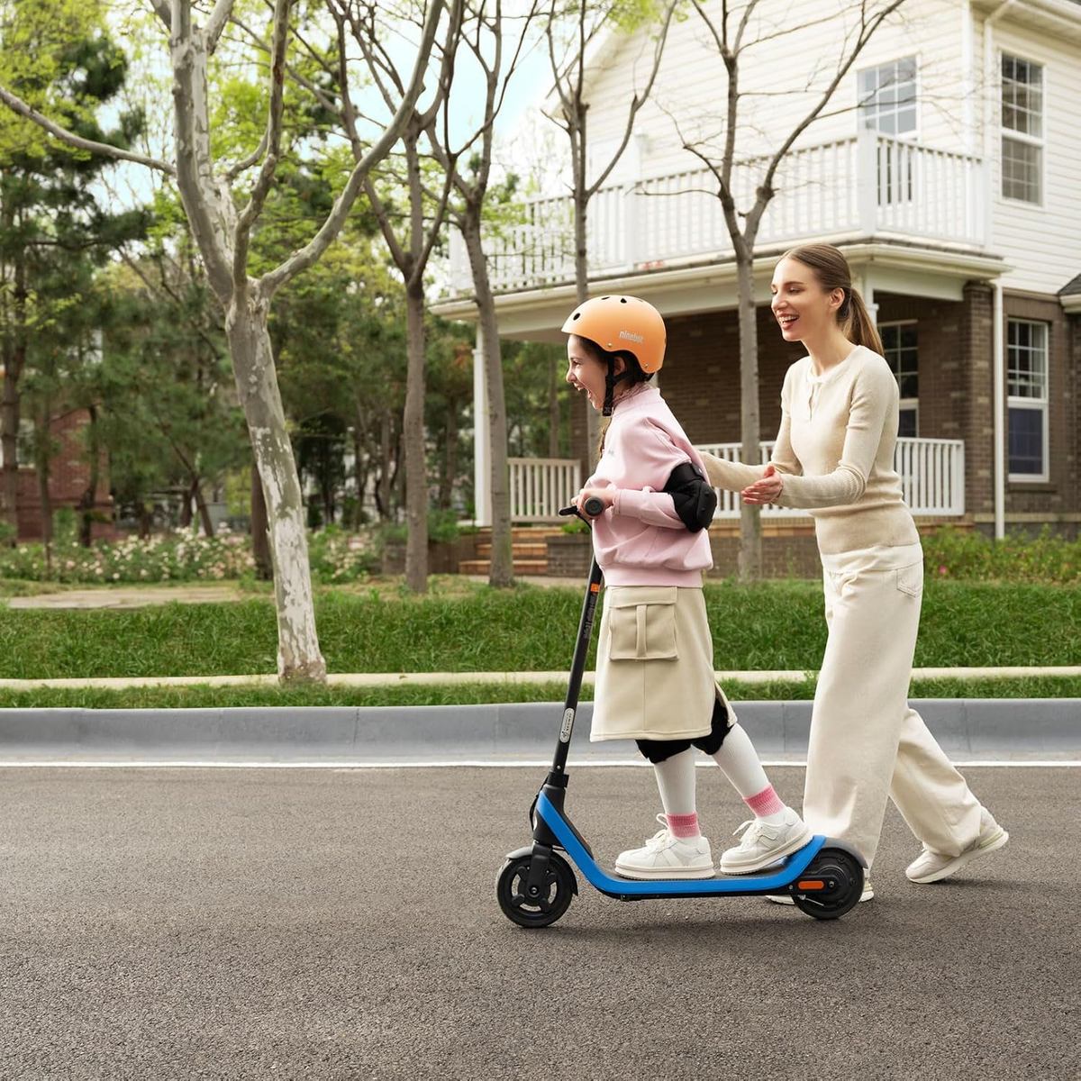Una ragazza va in scooter blu con una donna che cammina accanto. Sono in strada davanti a una casa.